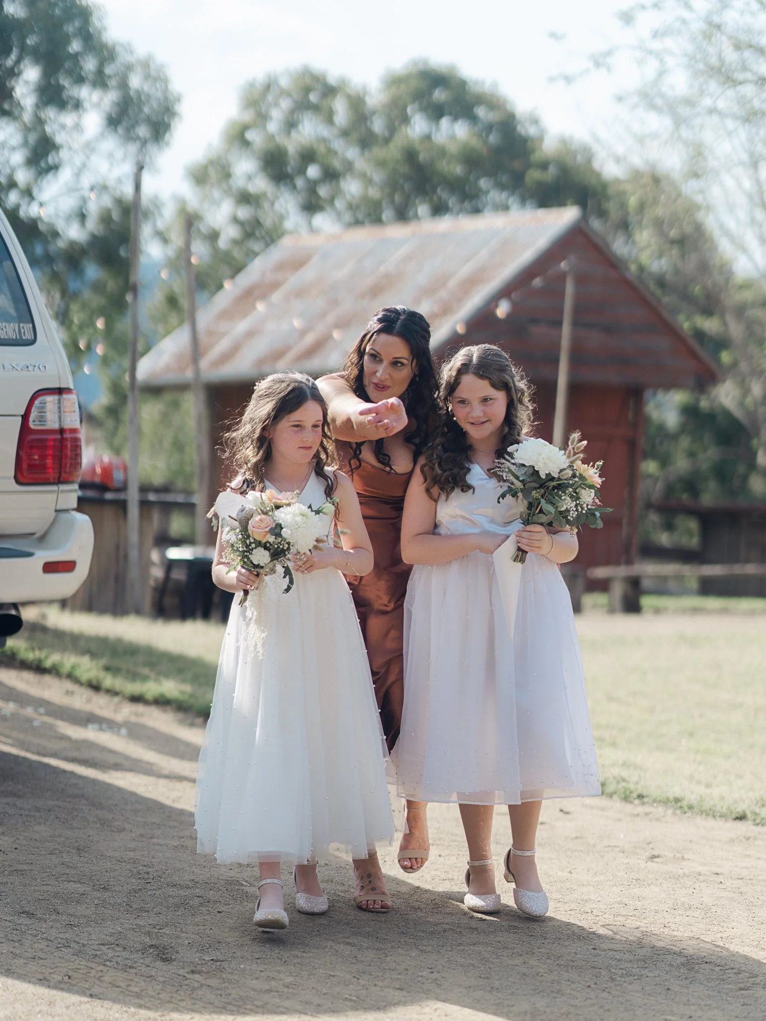 Hobart wedding photographer capturing A woman and two young girls walking outdoors, with the woman pointing, each girl holding a bouquet of flowers, on a sunny day near a rustic building.