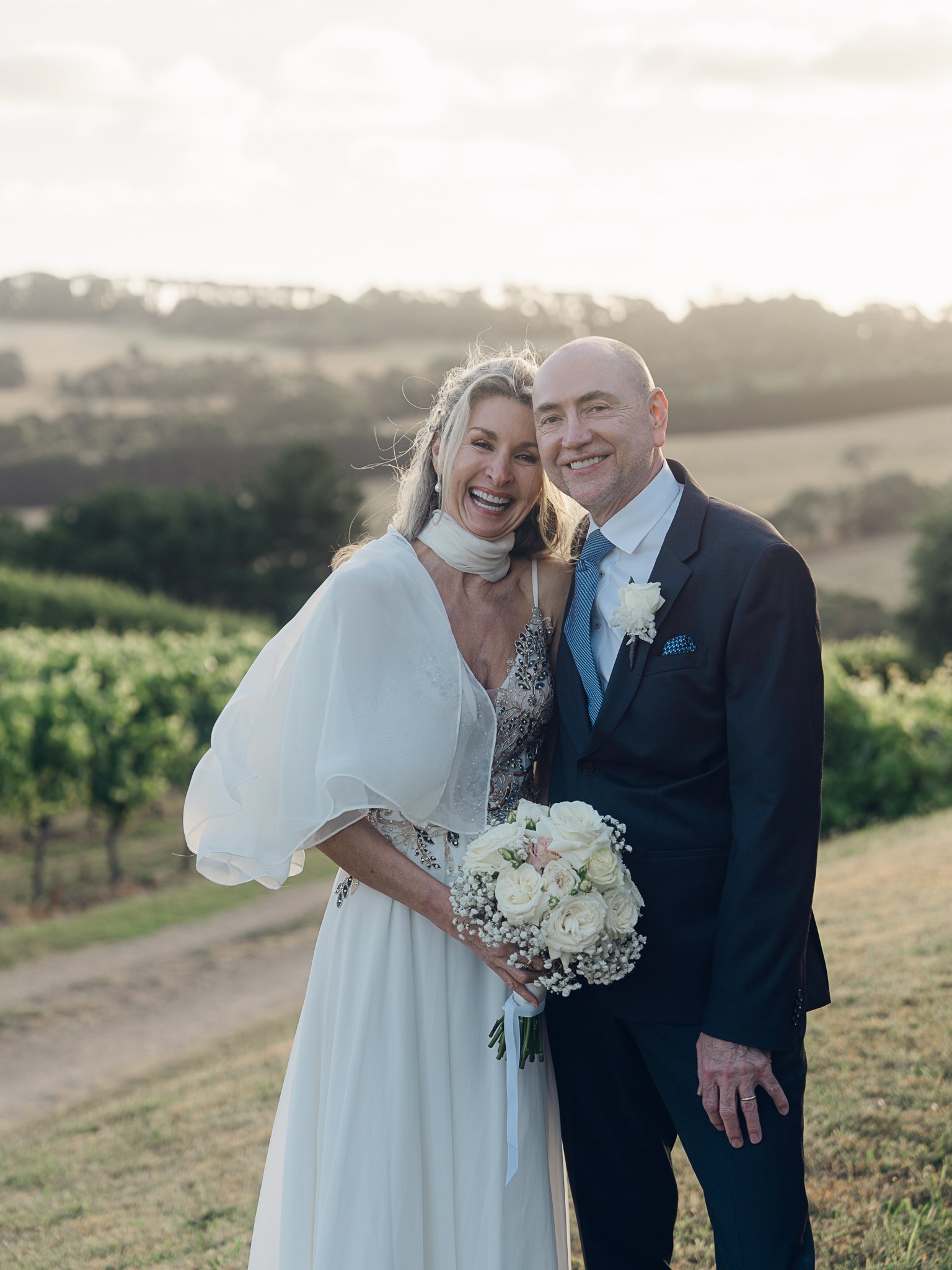 Happy bride and groom celebrating outdoors during their wedding, holding a bouquet of white roses, with a scenic countryside background