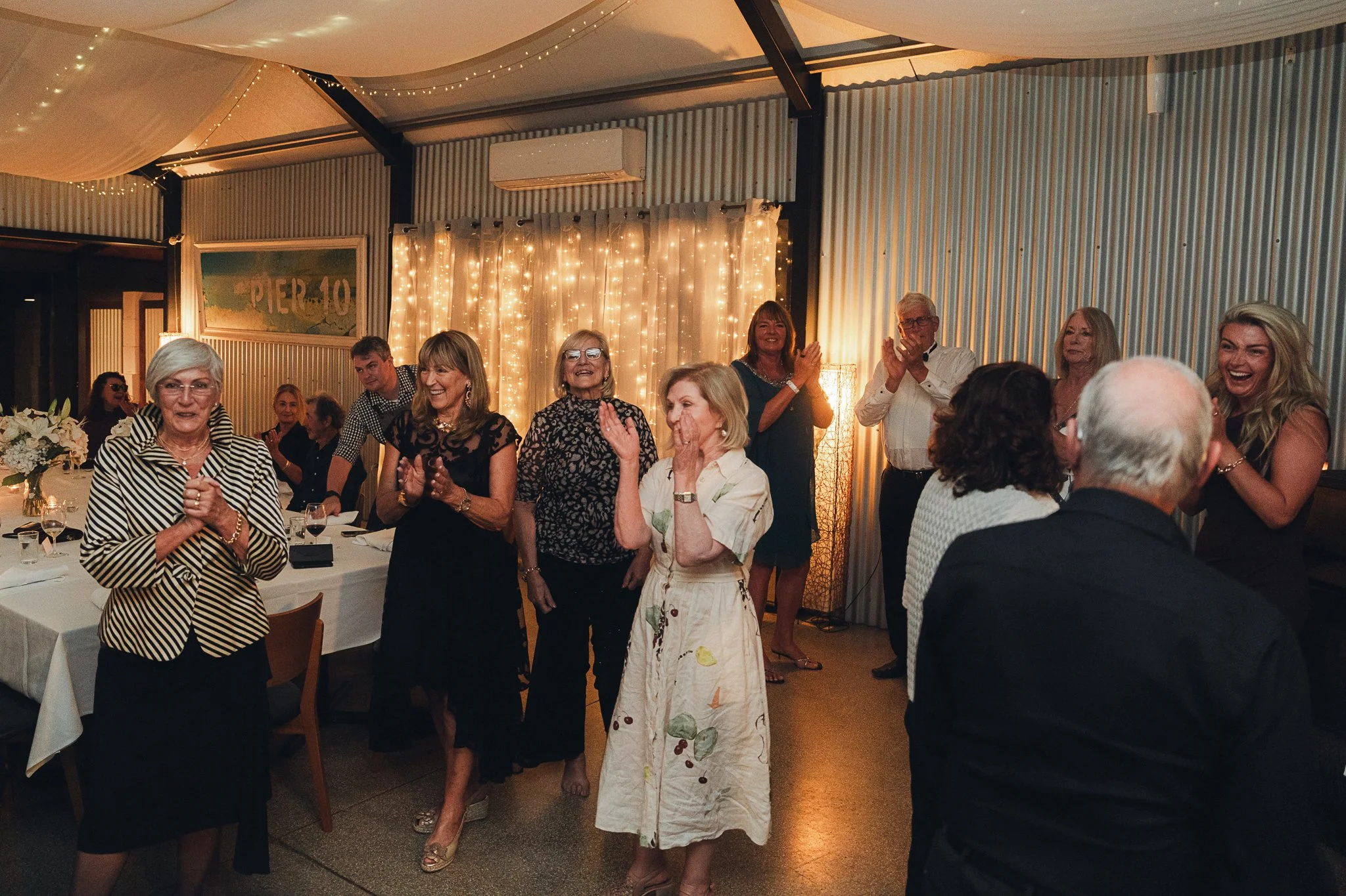 Group of people celebrating in a decorated indoor setting with fairy lights and draped fabric, clapping and smiling.