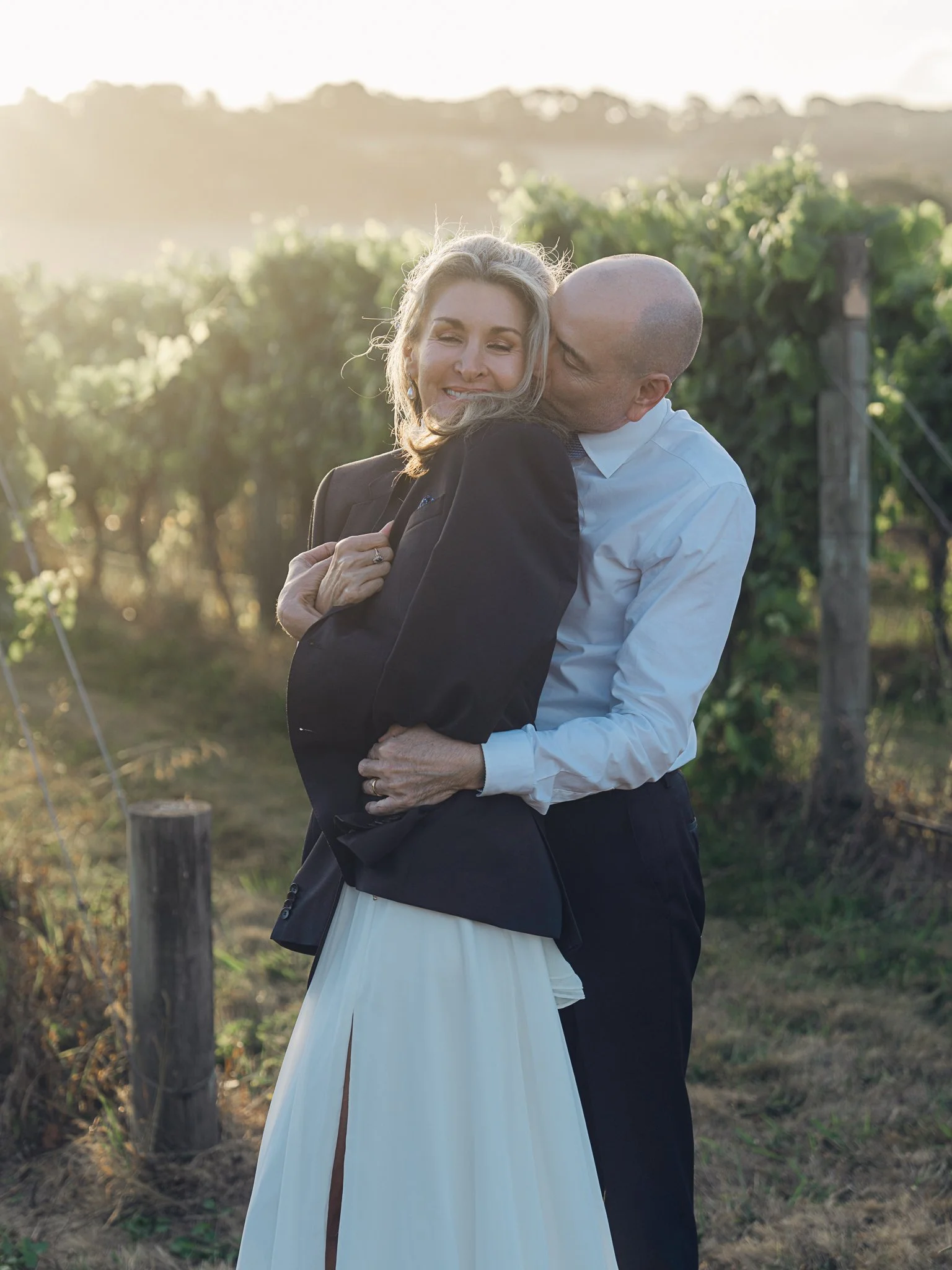 A couple hugging outdoors, with the man kissing the woman's cheek as they stand in a vineyard during sunset.