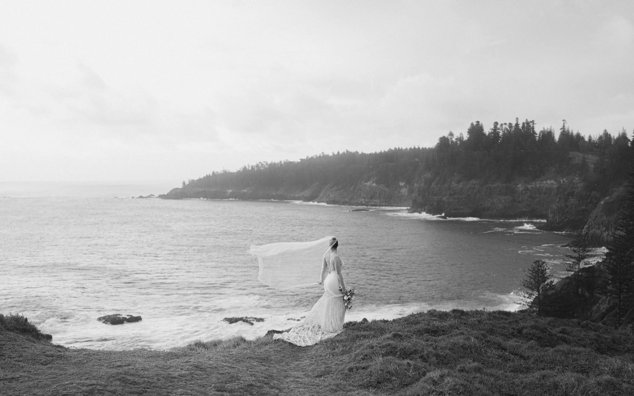 Hobart wedding photographer capturing a photo of a woman in a wedding dress standing on grassy cliffs overlooking the ocean, with a long veil flowing in the wind and holding a bouquet of flowers, with a coastline and trees in the background.