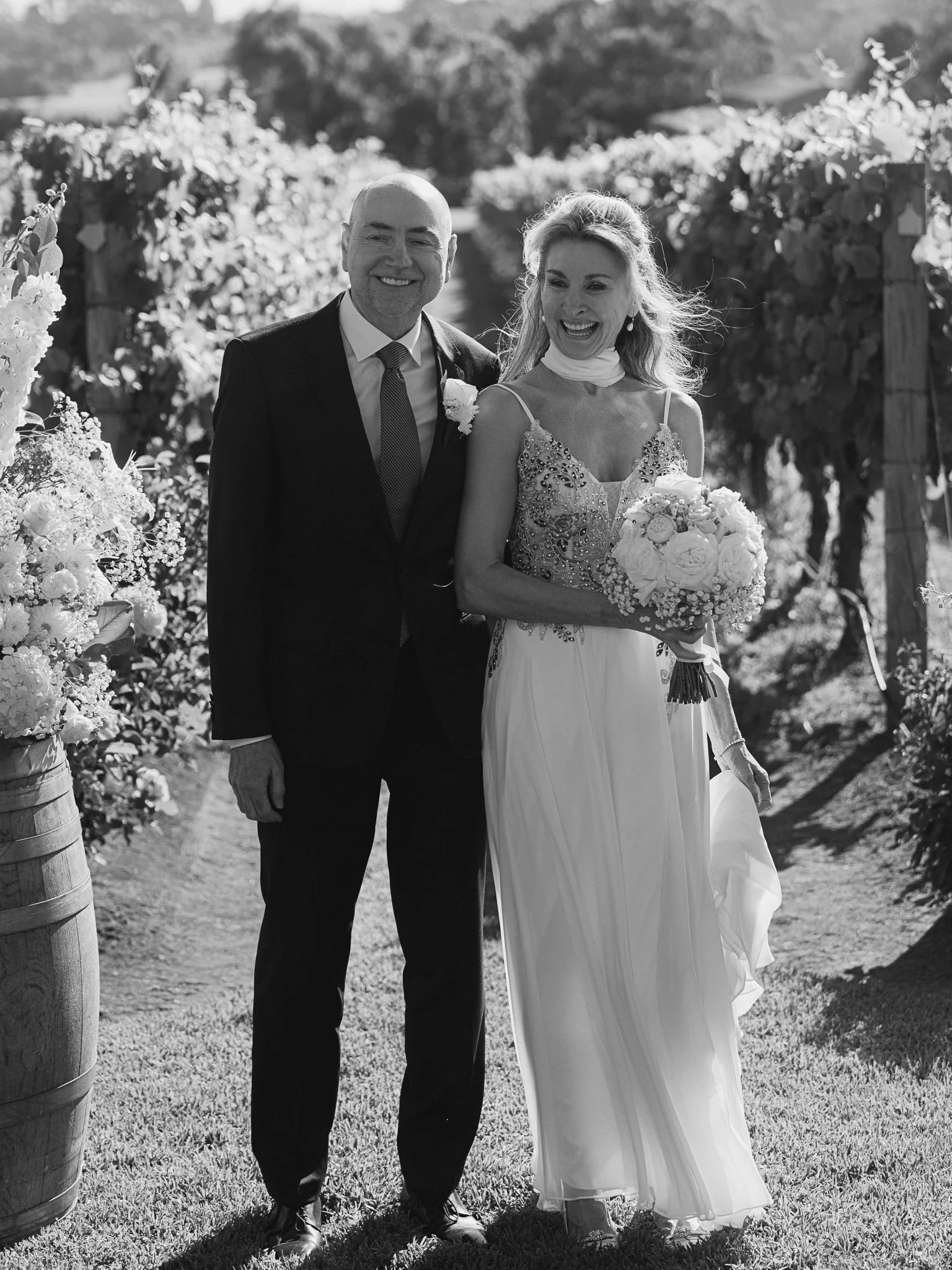 A newlywed couple walking outdoors in a vineyard, with the bride holding a bouquet of flowers and wearing a detailed dress, and the groom dressed in a suit, both smiling.