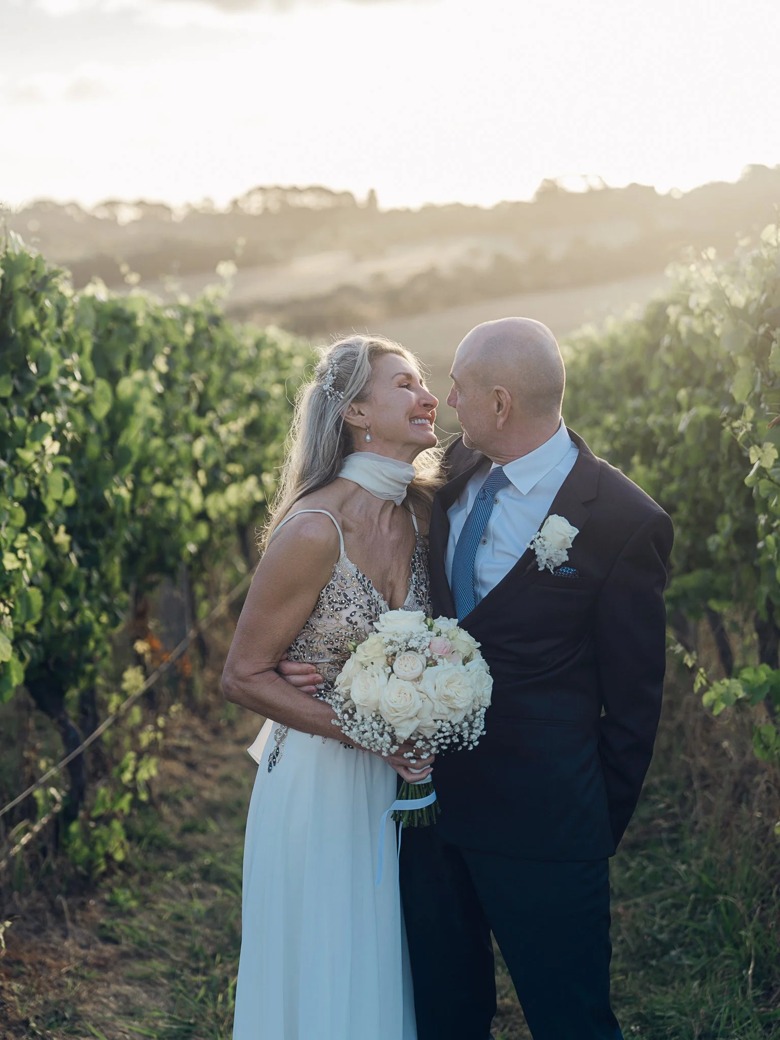 An elderly woman in a wedding dress smiling at an elderly man in a suit, standing in a vineyard during sunset, holding a bouquet of white roses.
