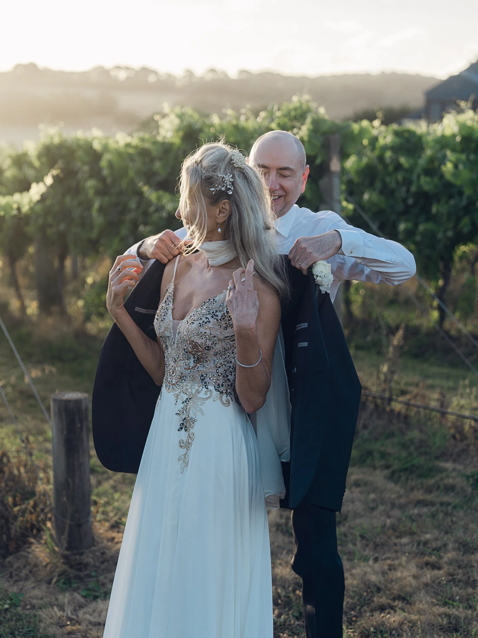 A couple in wedding attire embracing outdoors in a vineyard at sunset, smiling and sharing a joyful moment.