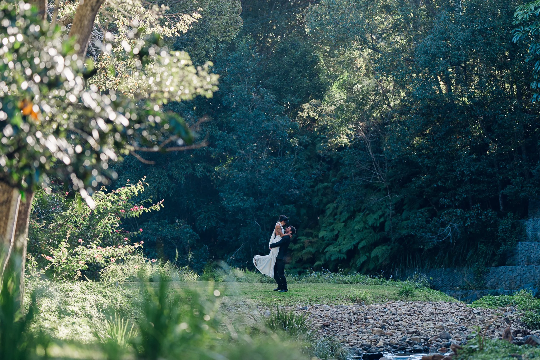 A bride and groom dancing outdoors in a lush, green forest setting with sunlight filtering through trees.