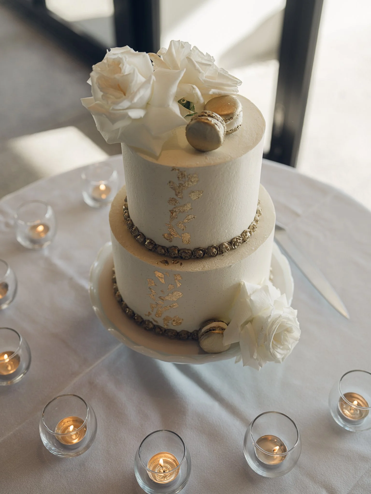A two-tiered white wedding cake decorated with white flowers, macarons, gold leaf accents, and small edible pearls, surrounded by small lit candles.