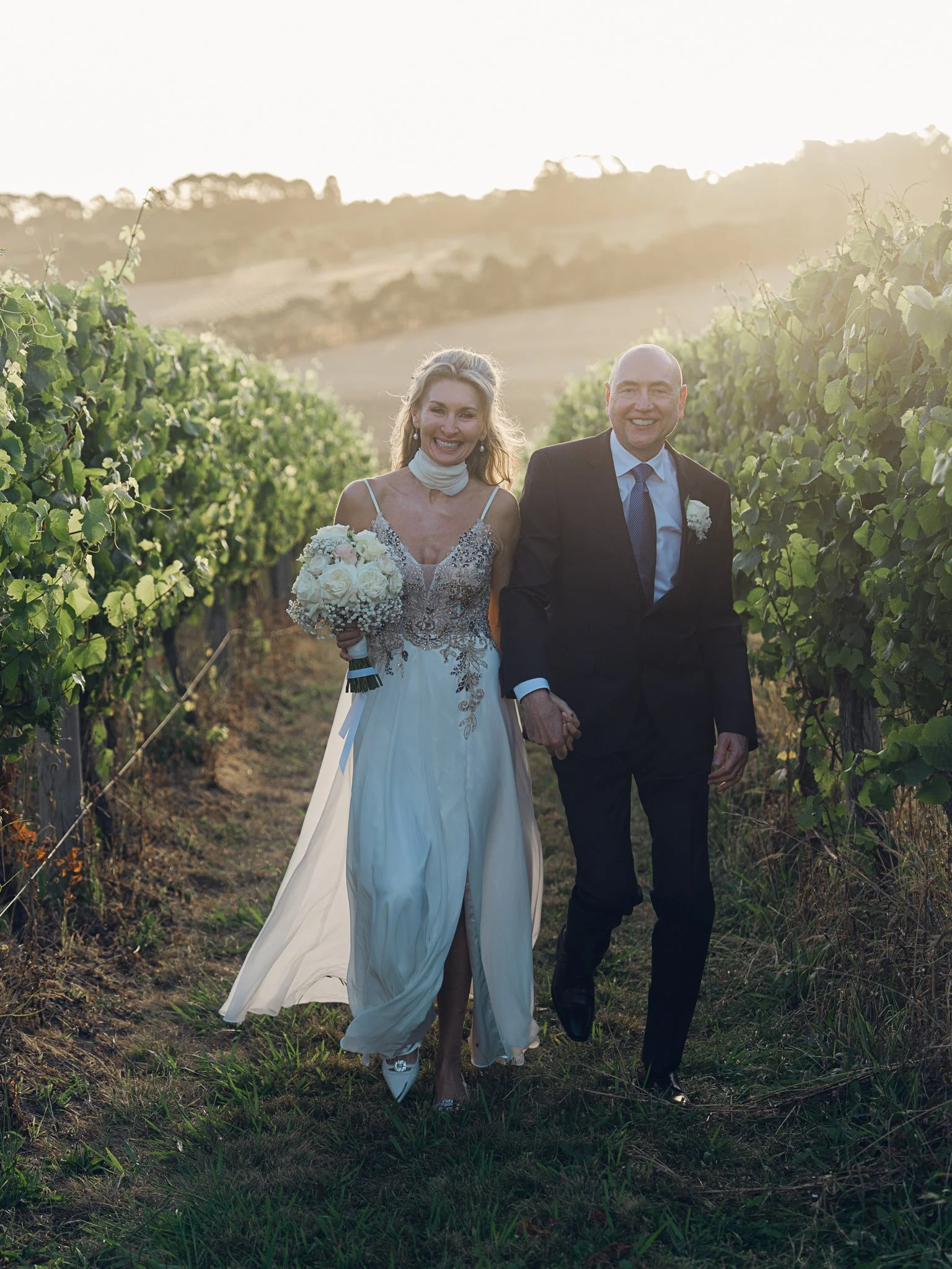 A bride and groom walking hand in hand through a vineyard at sunset, smiling happily.