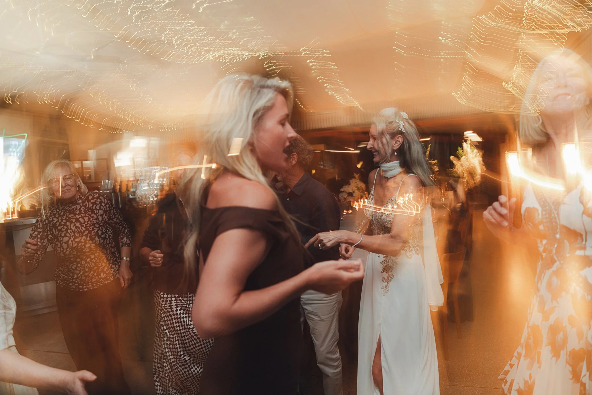 People dancing and socializing at a celebration or wedding reception in a warmly lit indoor venue with motion blur.