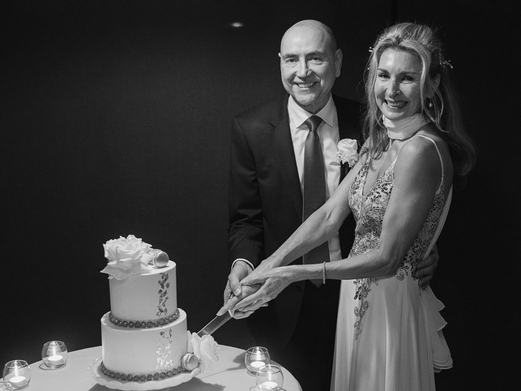 A happy couple, a bride and groom, cutting their wedding cake together at a wedding reception. The groom is dressed in a suit and tie, and the bride is in a formal gown with intricate embroidery. The cake has two tiers, decorated with flowers and a cake topper, and is surrounded by small lit candles.