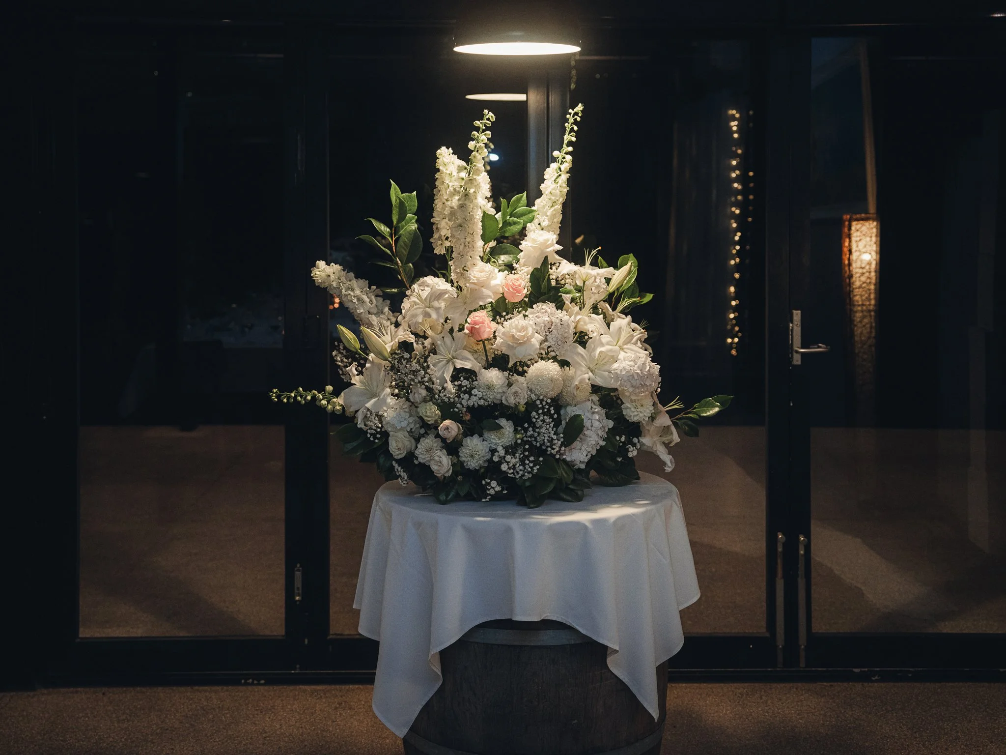 A floral arrangement with white and light pink flowers on a table with a white tablecloth, illuminated by a ceiling light, against a dark background.