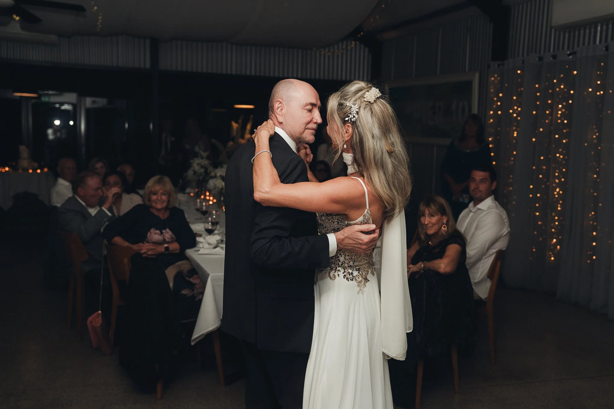 An older man and woman dance closely at a wedding reception, with guests seated at tables and fairy lights in the background.