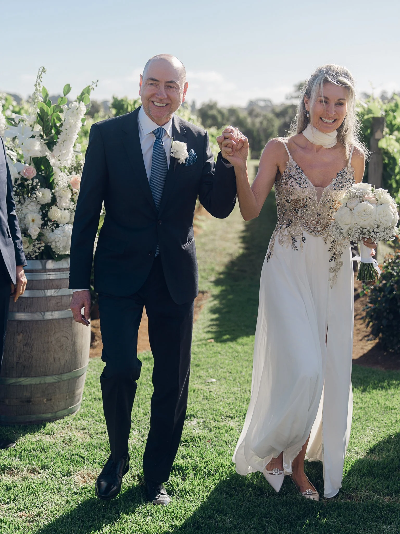 A bride and groom holding hands and smiling during a wedding ceremony outdoors on a sunny day, with flower arrangements around them.