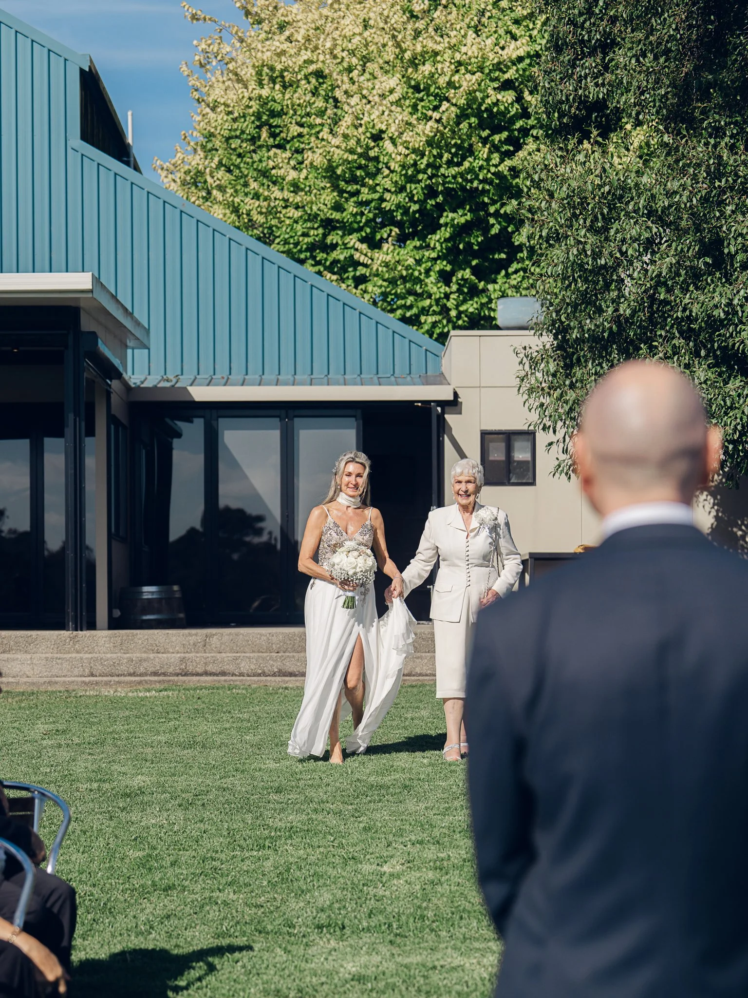 A bride in a white wedding dress holding a bouquet of white flowers is walking on a grassy area, accompanied by an older woman in white, as she approaches a man in a dark suit at an outdoor wedding ceremony.