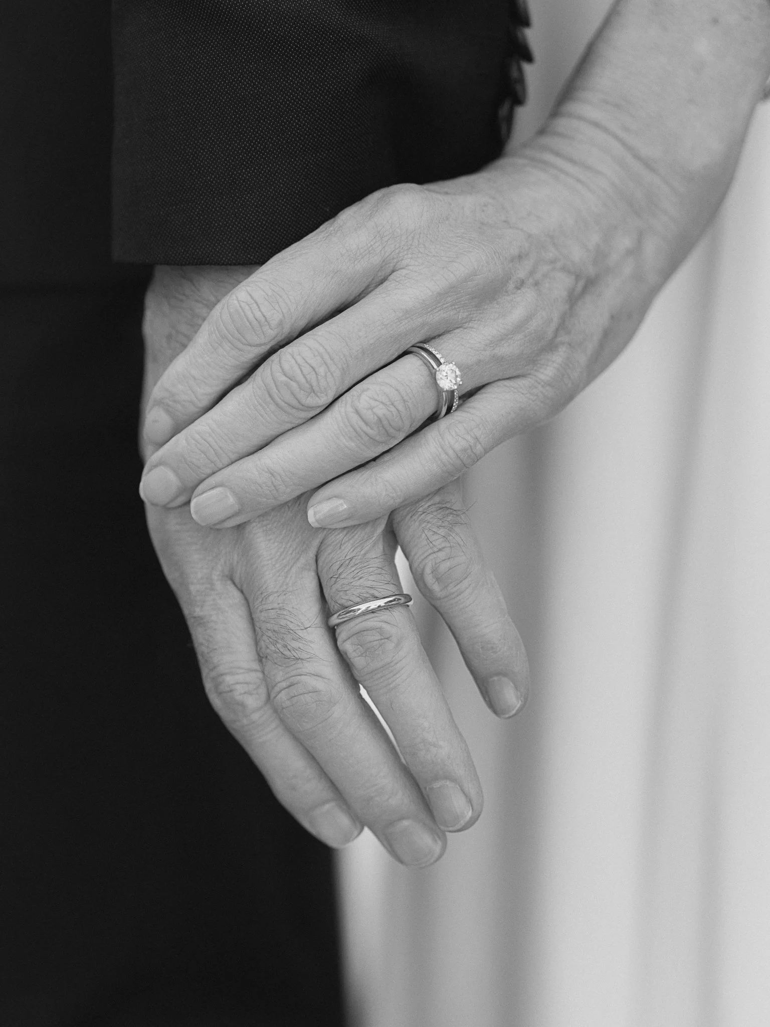 Close-up of a couple's hands with wedding rings, with one hand resting on top of the other, in black and white.