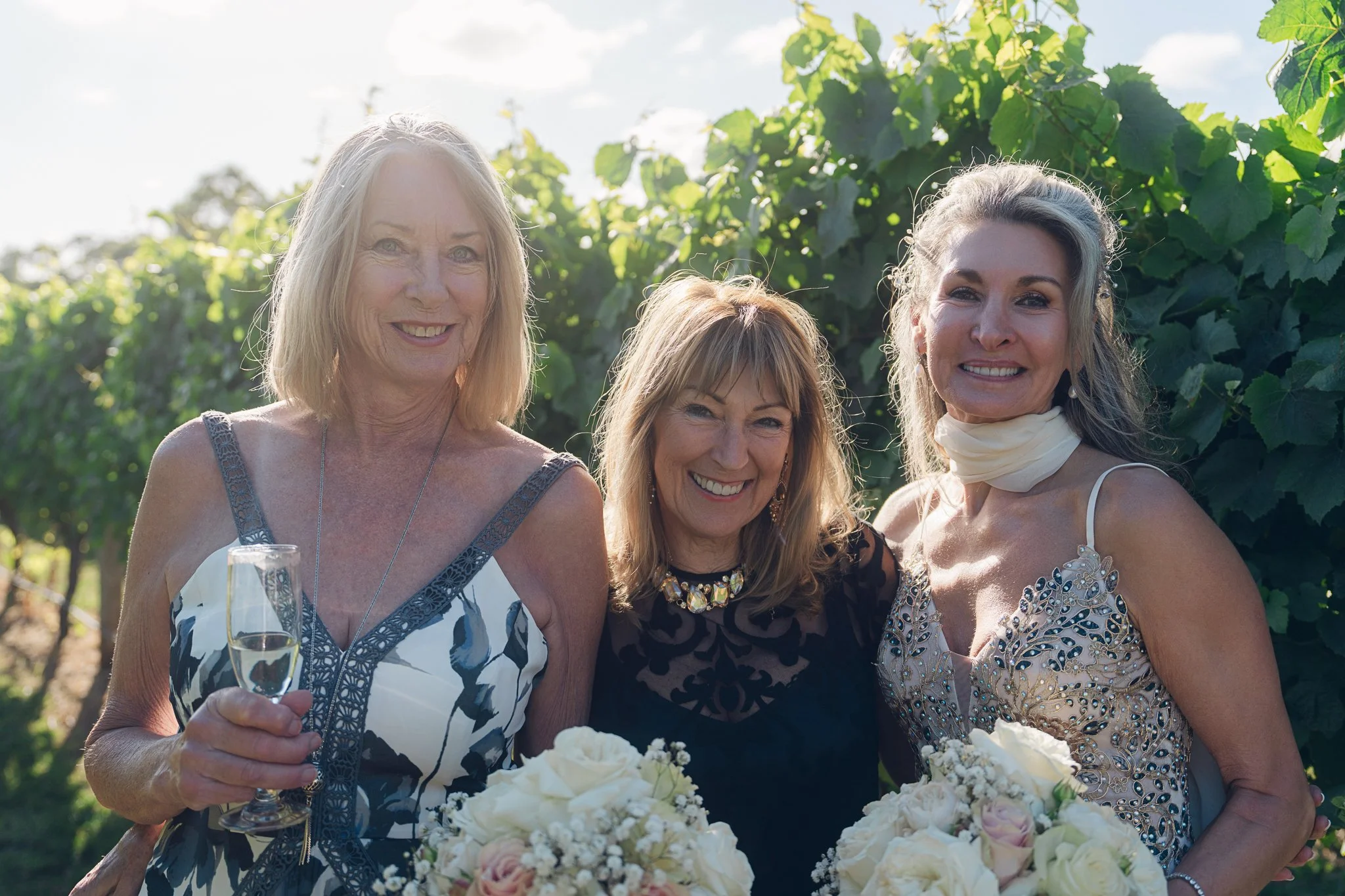 Three women smiling outdoors during a celebration, holding bouquets of white flowers, with vineyard foliage in the background.