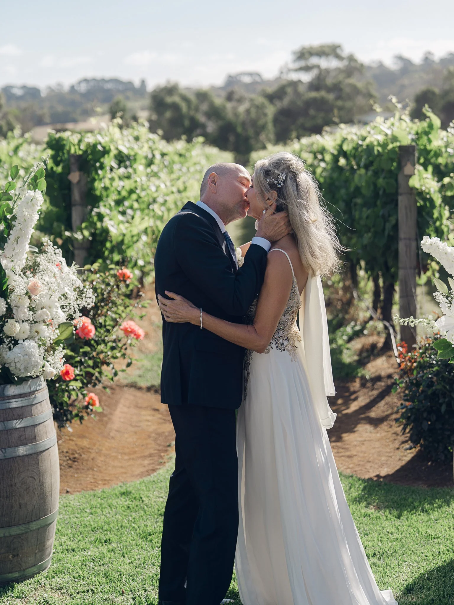 A couple in wedding attire sharing a kiss in a vineyard with greenery and flowers.