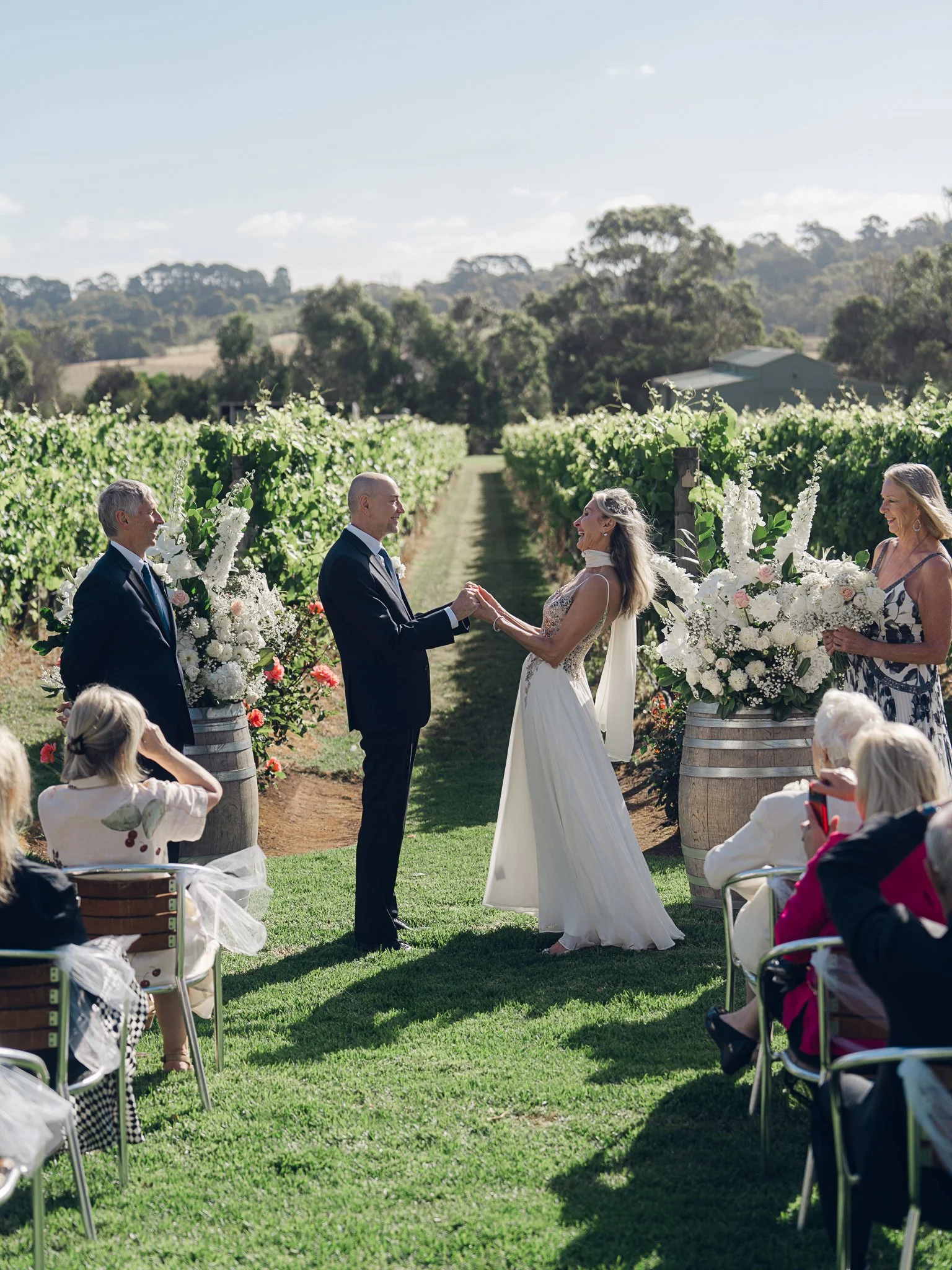 A wedding ceremony outdoors on a sunny day in a vineyard with a bride and groom holding hands, surrounded by floral arrangements, guests seated, and green vineyard rows in the background.