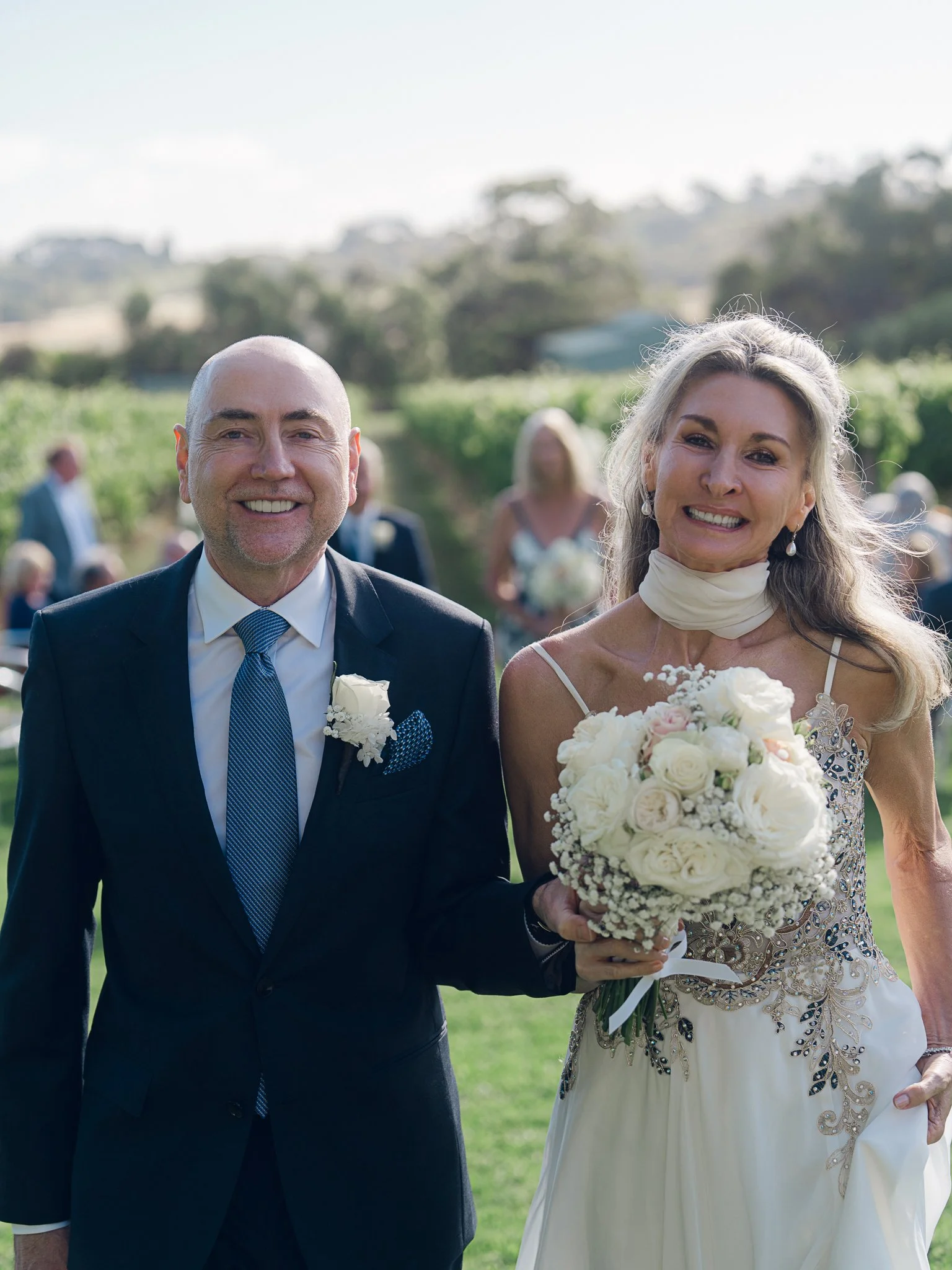 A joyful bride and groom smiling outdoors on their wedding day, the bride holding a bouquet of white roses and wearing a strapless wedding dress, the groom in a dark suit with a boutonniere, with blurred wedding guests in the background.