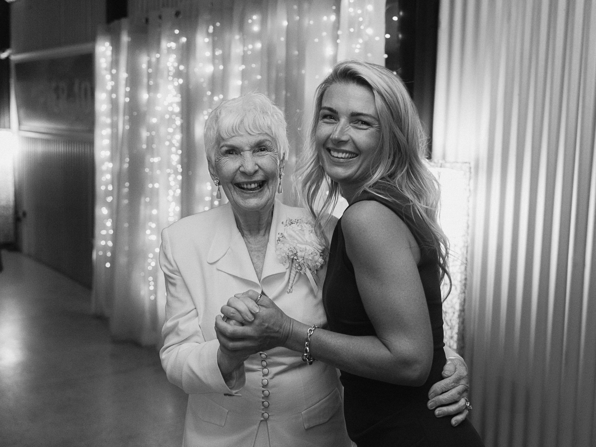An elderly woman and a young woman happily dancing together at an indoor celebration, with string lights in the background.