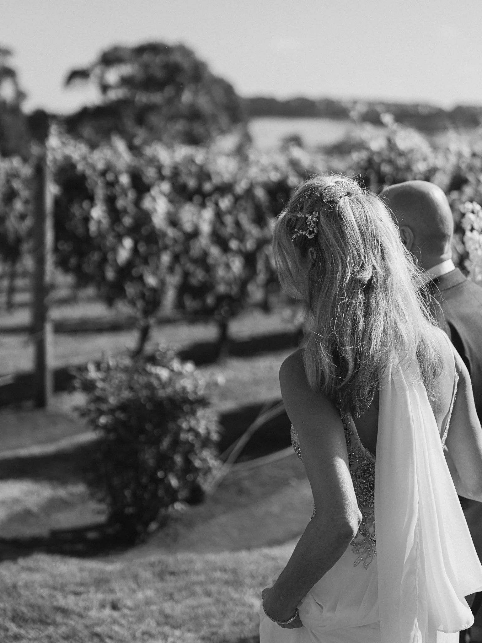 Black and white photo of a bride and groom walking outdoors with trees and a fence in the background.