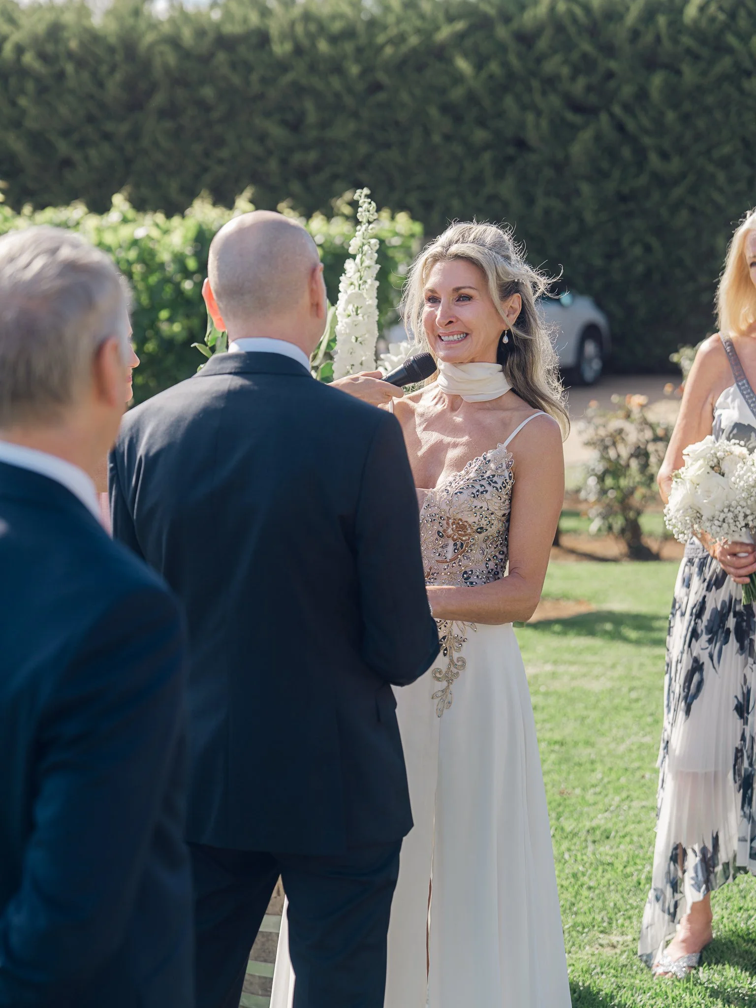 A woman in a wedding dress smiling as she speaks into a microphone during a wedding ceremony outdoors, with a man in a suit facing her and other guests nearby.