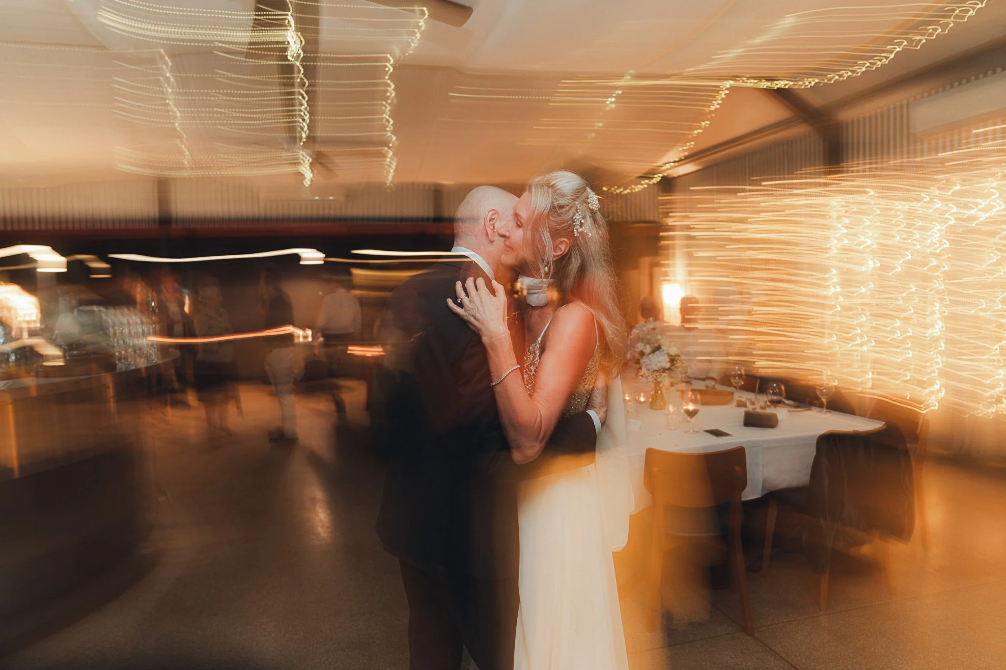 A bride and groom are dancing closely at their wedding reception in a dimly lit room with string lights, causing a motion blur effect.