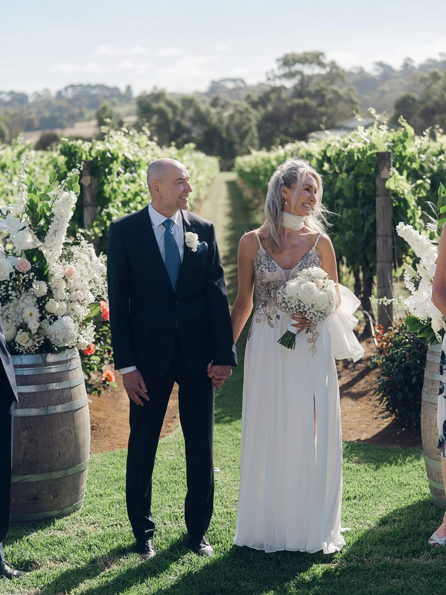 A bride and groom holding hands during their outdoor wedding ceremony in a vineyard, surrounded by large flower arrangements and lush green grapevines, with a scenic hillside in the background.