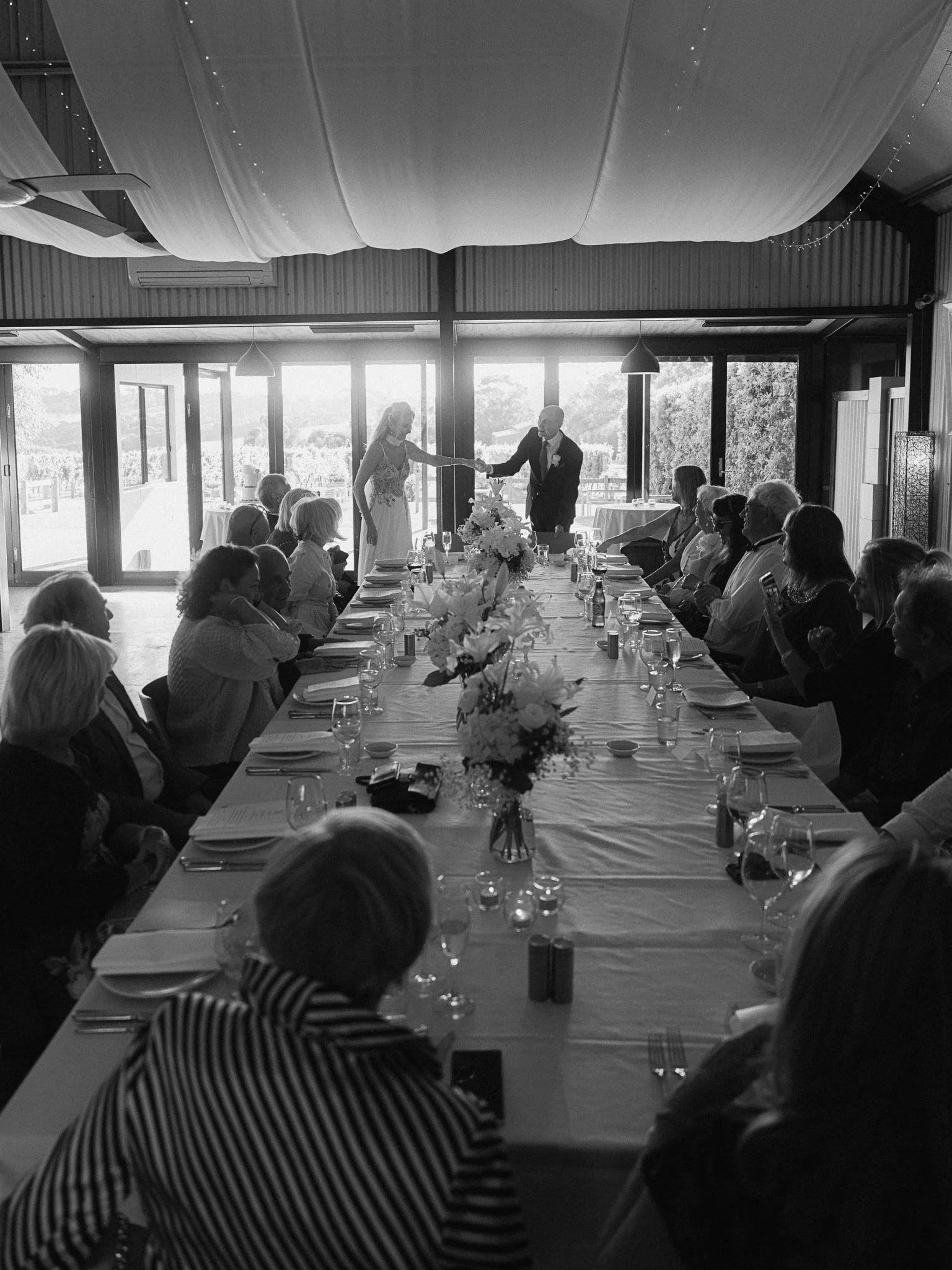 Black and white photo of a wedding reception with a couple dancing at the head of a long banquet table surrounded by seated guests, with large windows and sliding doors in the background.