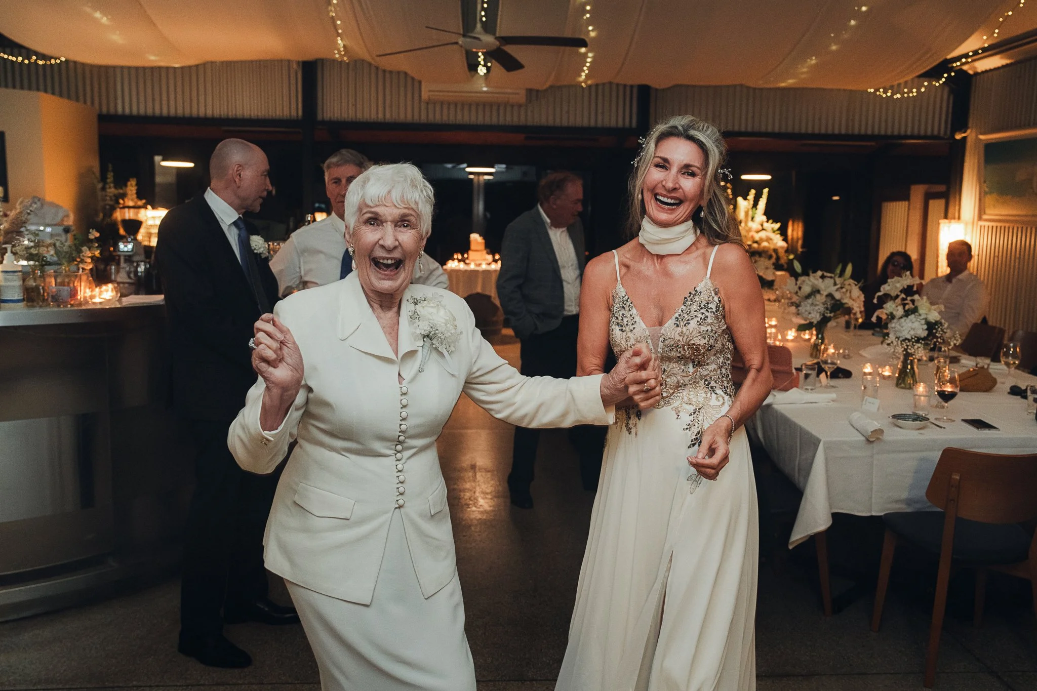 Two women, an elderly woman and a middle-aged woman, smiling and dancing at a wedding reception. The elderly woman is wearing a white suit, while the middle-aged woman is in a cream-colored dress with floral embroidery. The background shows a decorated reception hall with tables, flowers, and guests.