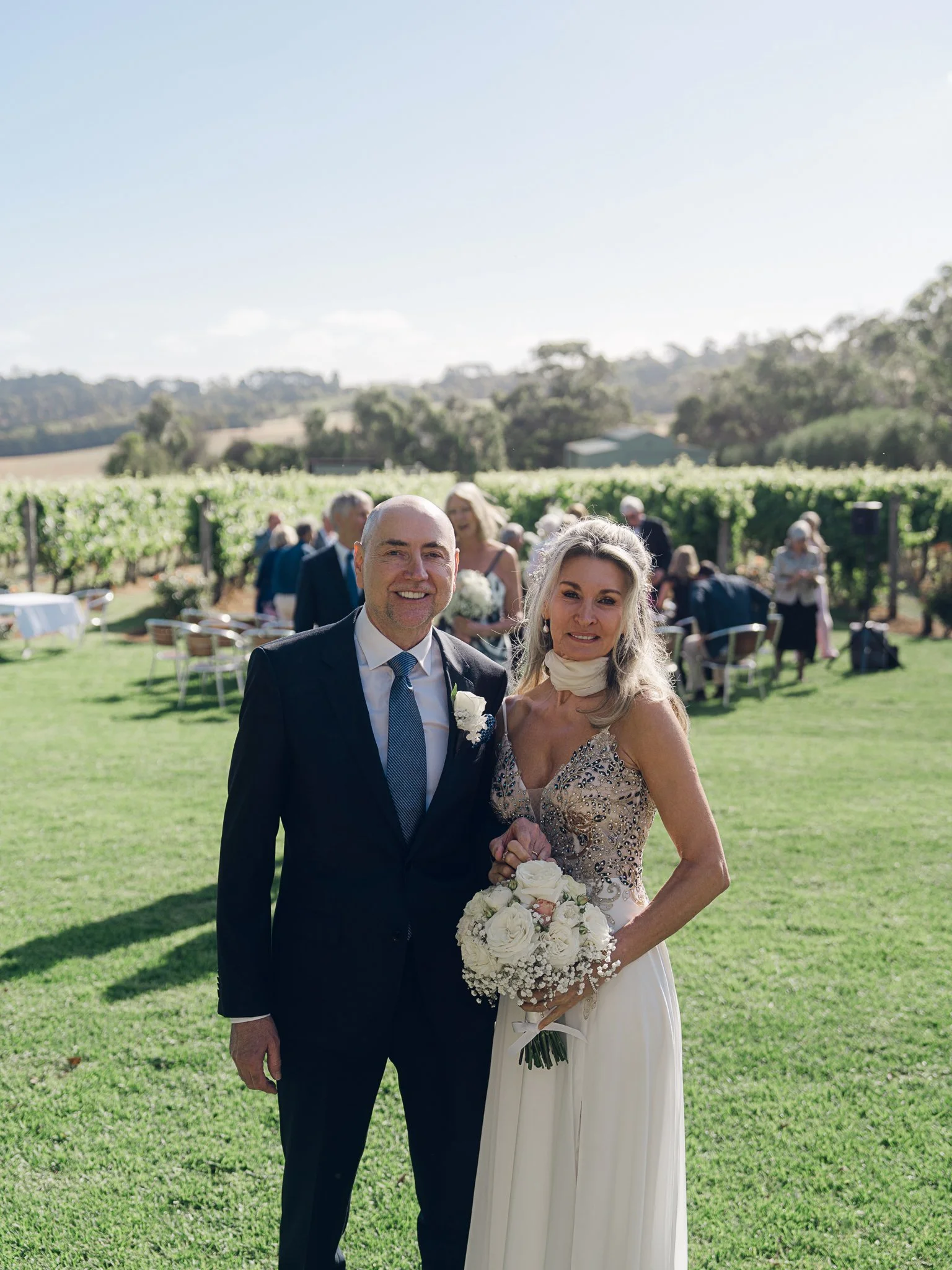 A bride and groom posing outdoors at a wedding ceremony in a vineyard, with guests in the background under a clear blue sky.