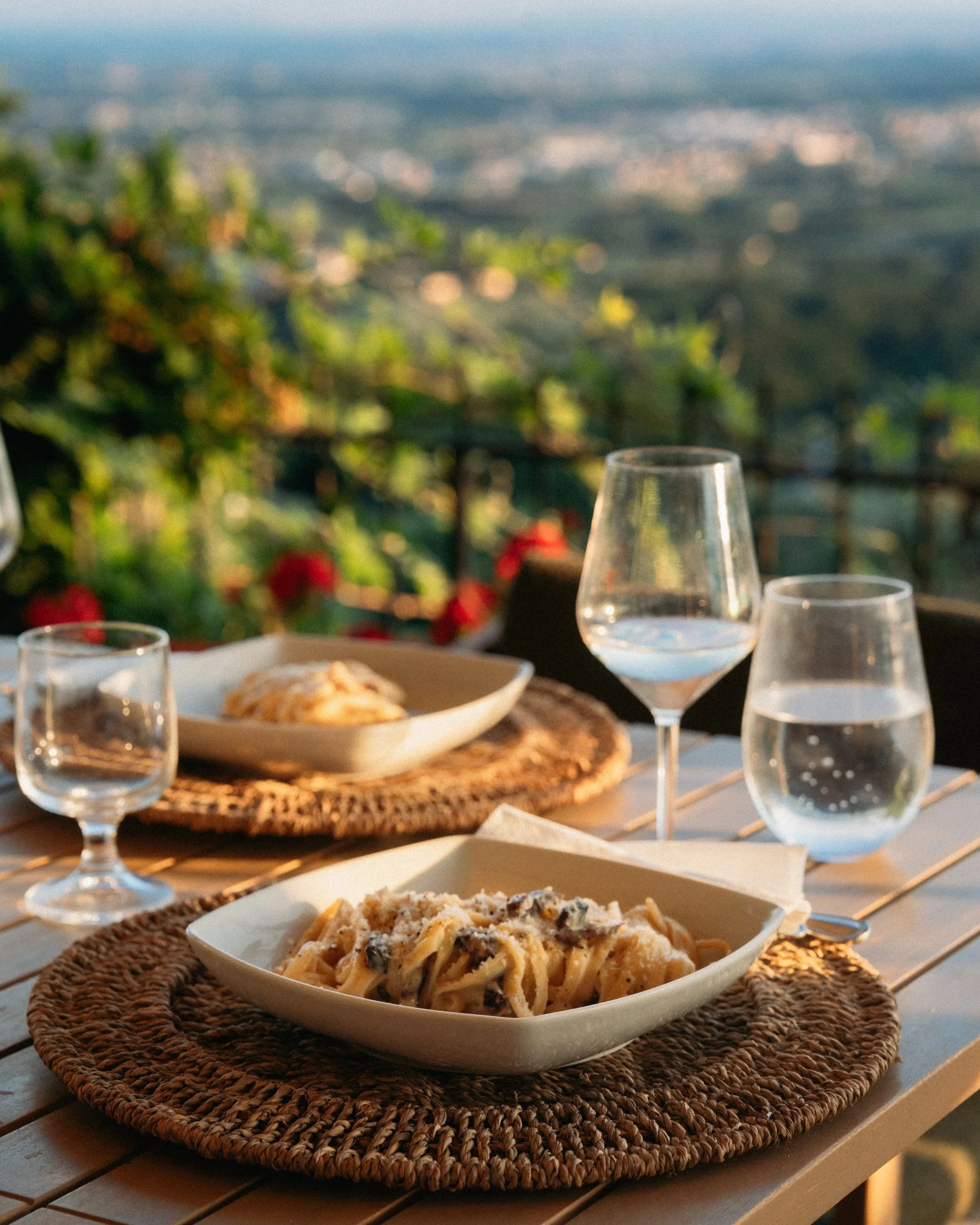 Close-up of a white oval dish filled with spaghetti topped with a creamy sauce and black pepper, placed on a woven placemat on a table. In the background, there are several wine glasses, some filled with water, with a scenic view of green hills and a