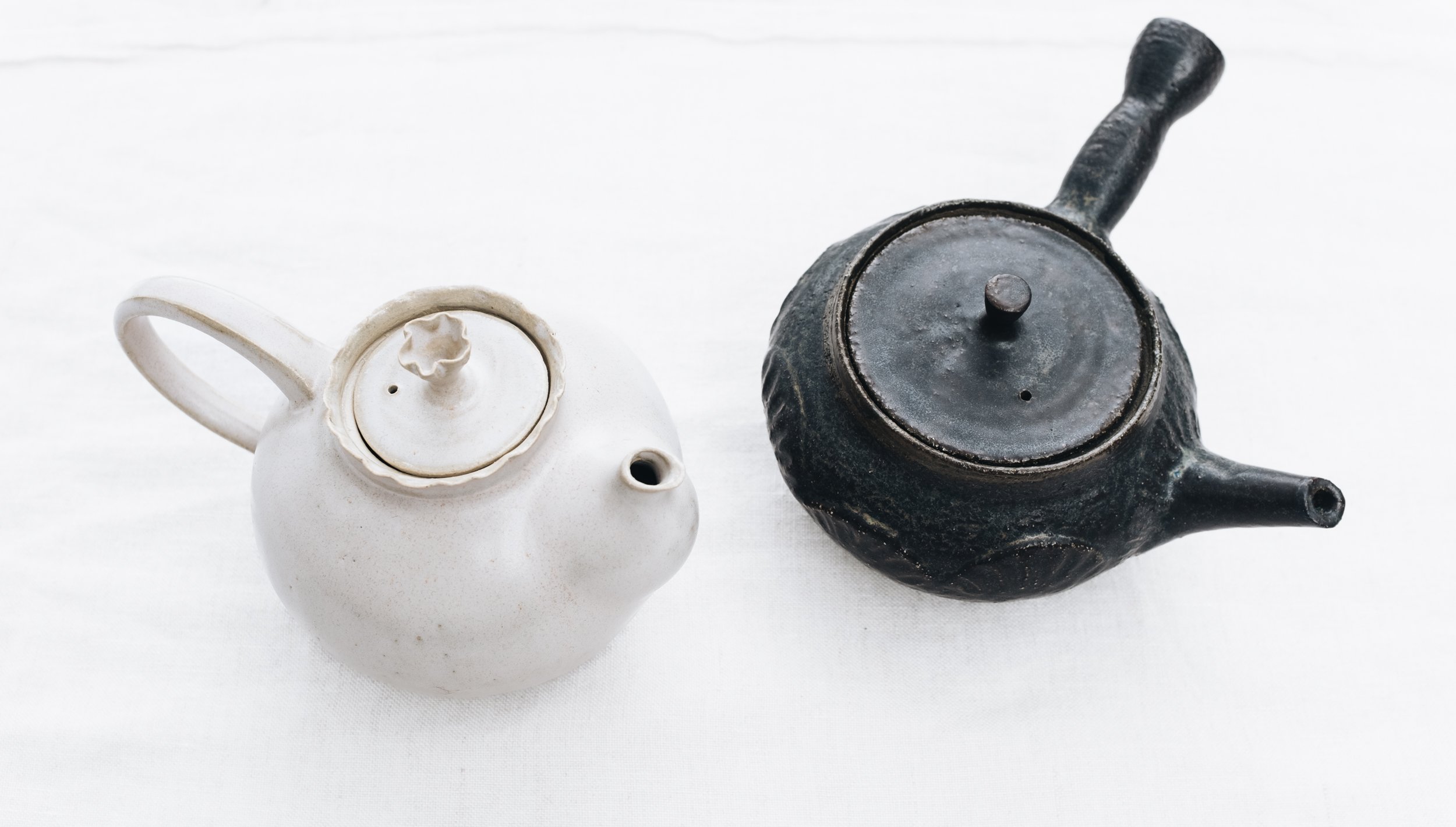 Overhead view of a white ceramic teapot and a black ceramic teapot, both with unique designs, on a white surface.