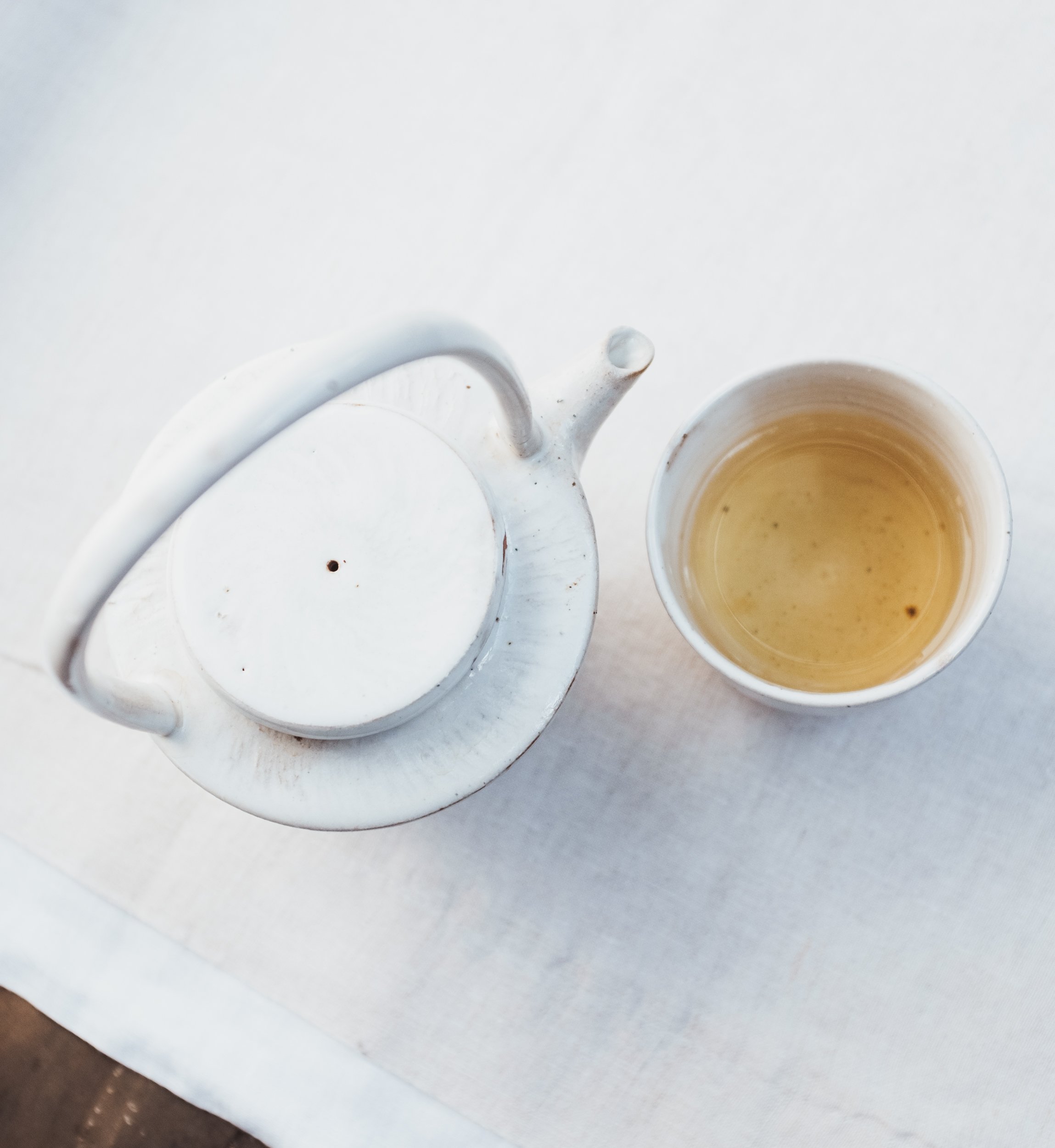 Overhead view of a white ceramic teapot and a cup of tea on a light surface.