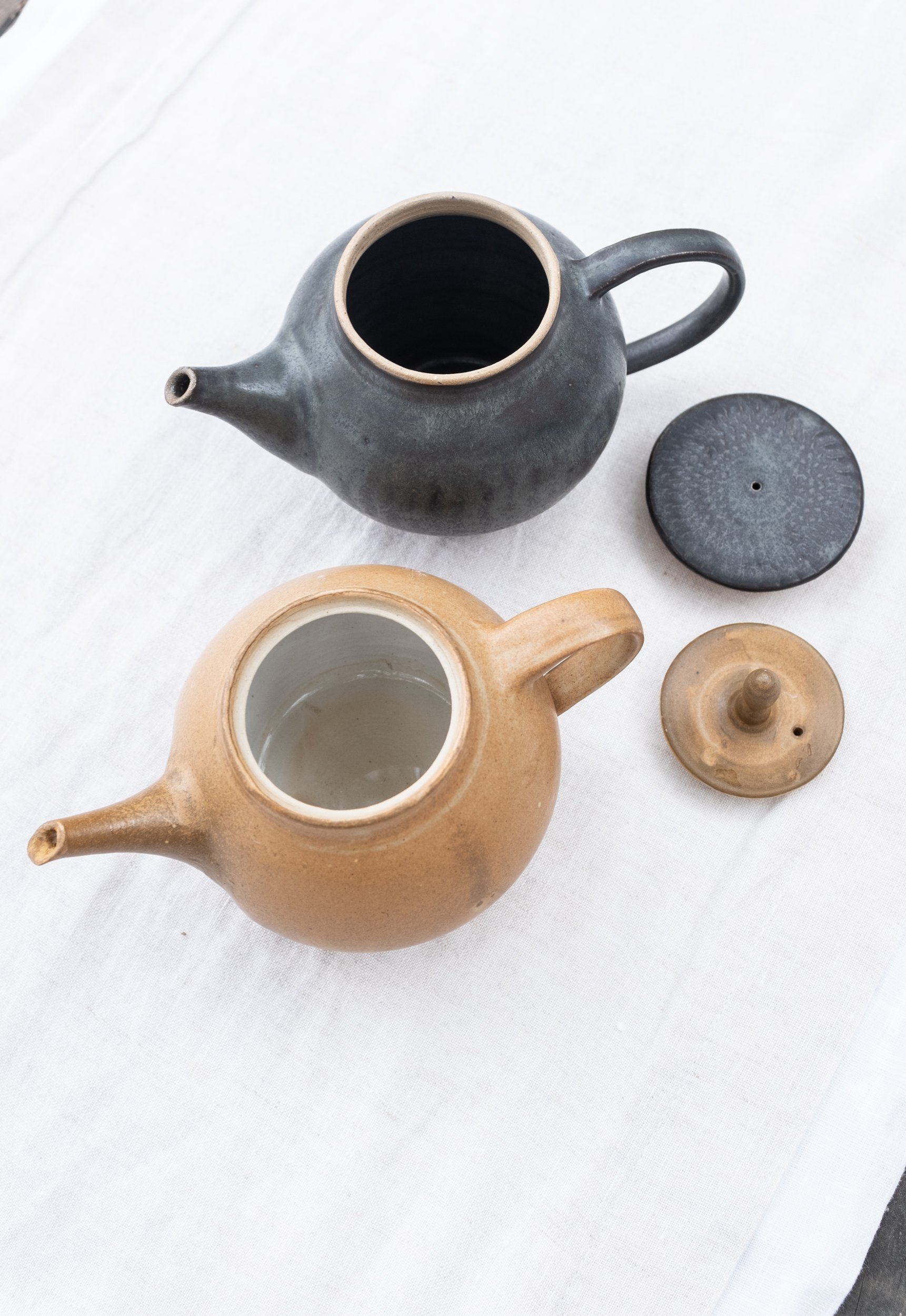 Two ceramic teapots with their lids off, one black and one tan, on a white fabric surface.
