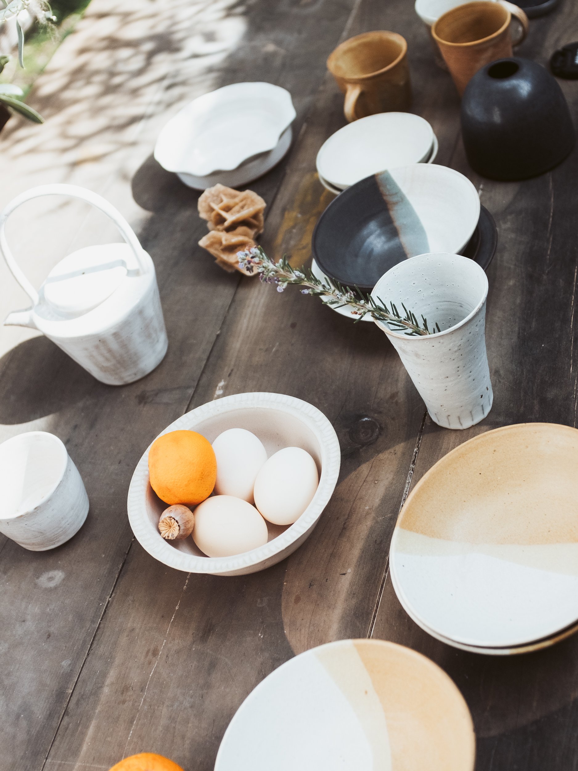 A rustic wooden table with ceramic bowls and cups, some containing eggs, an orange, a clove of garlic, and a small branch with flowers. There's also a white teapot and several empty ceramic dishes.