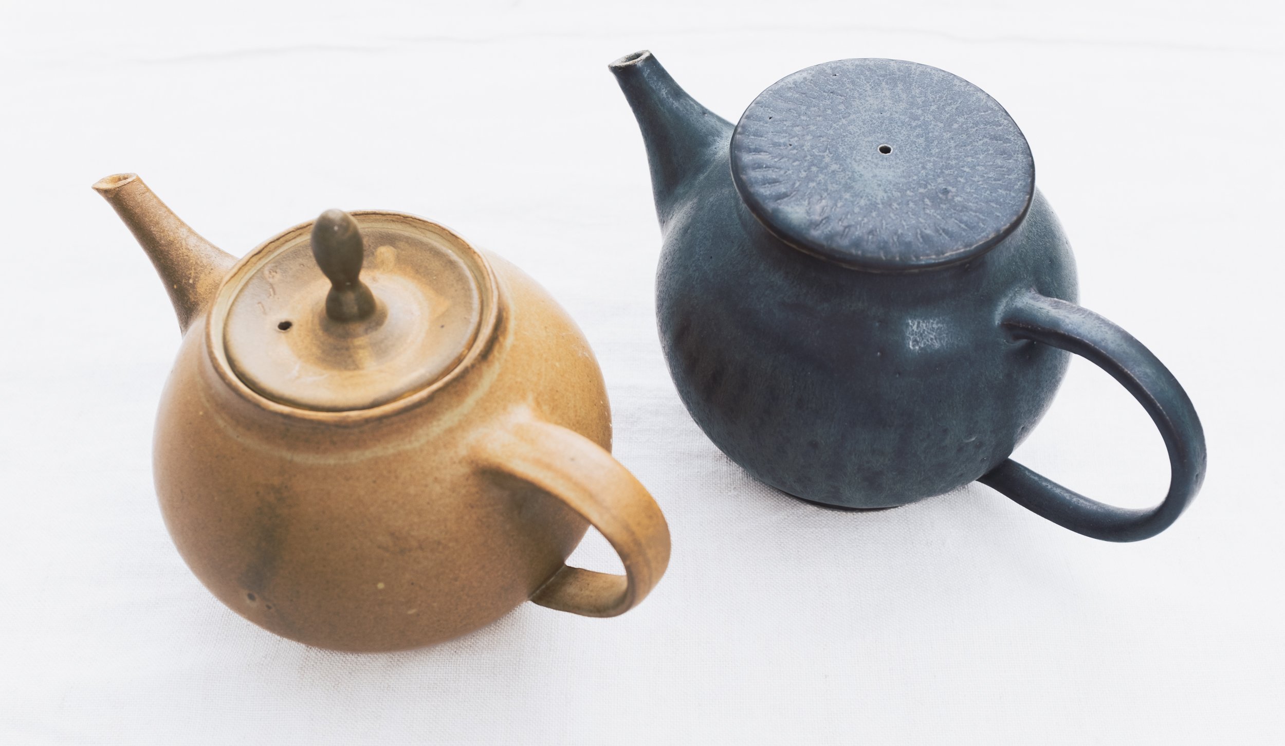 Two ceramic teapots, one brown and one blue, on a white surface.