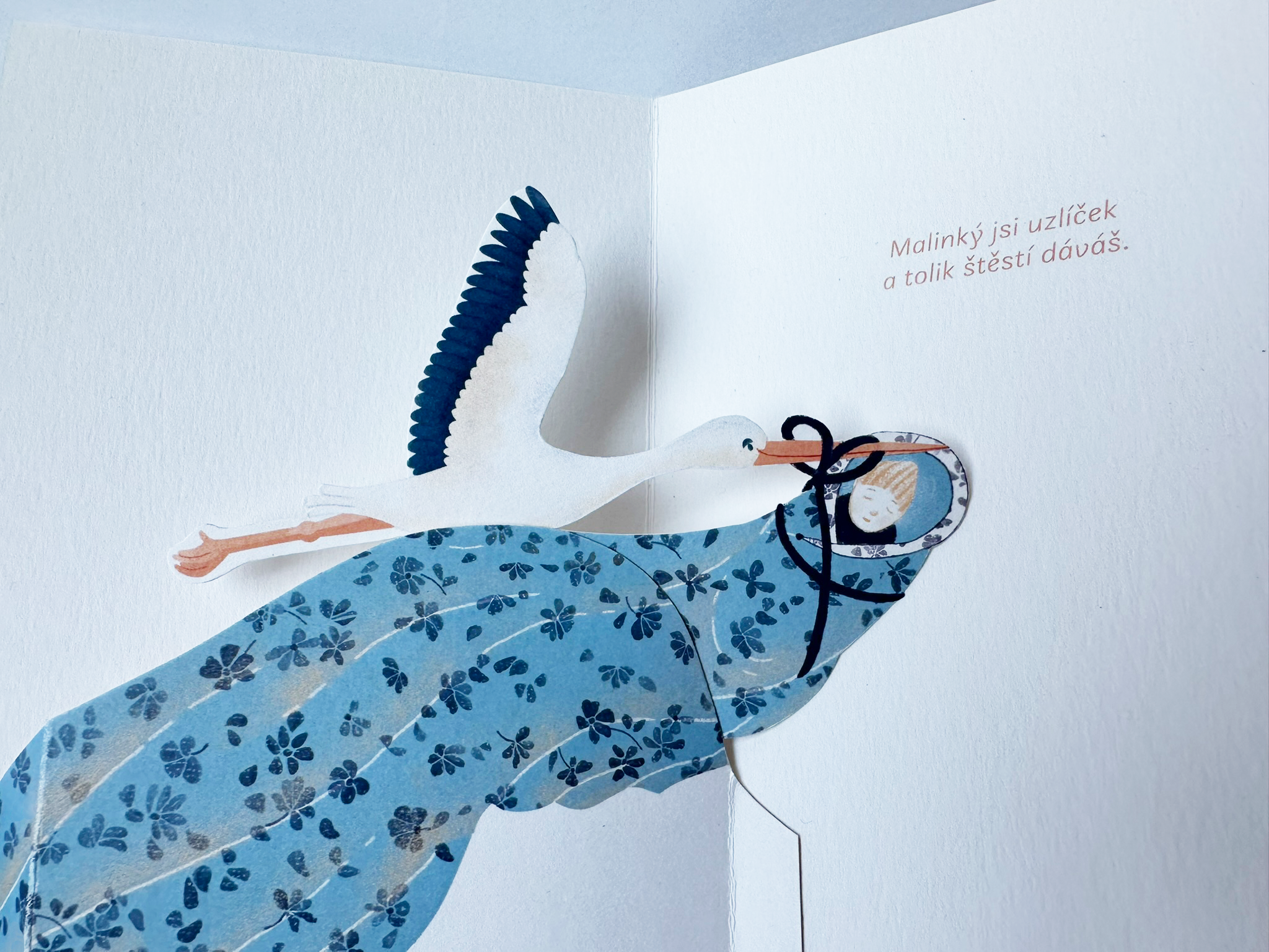 Pop-up ilustrovaný čáp nesoucí miminko zabalené do modré deky s květinovým vzorem a popiskem  „Malinký jsi uzlíček a tolik štěstí dáváš “