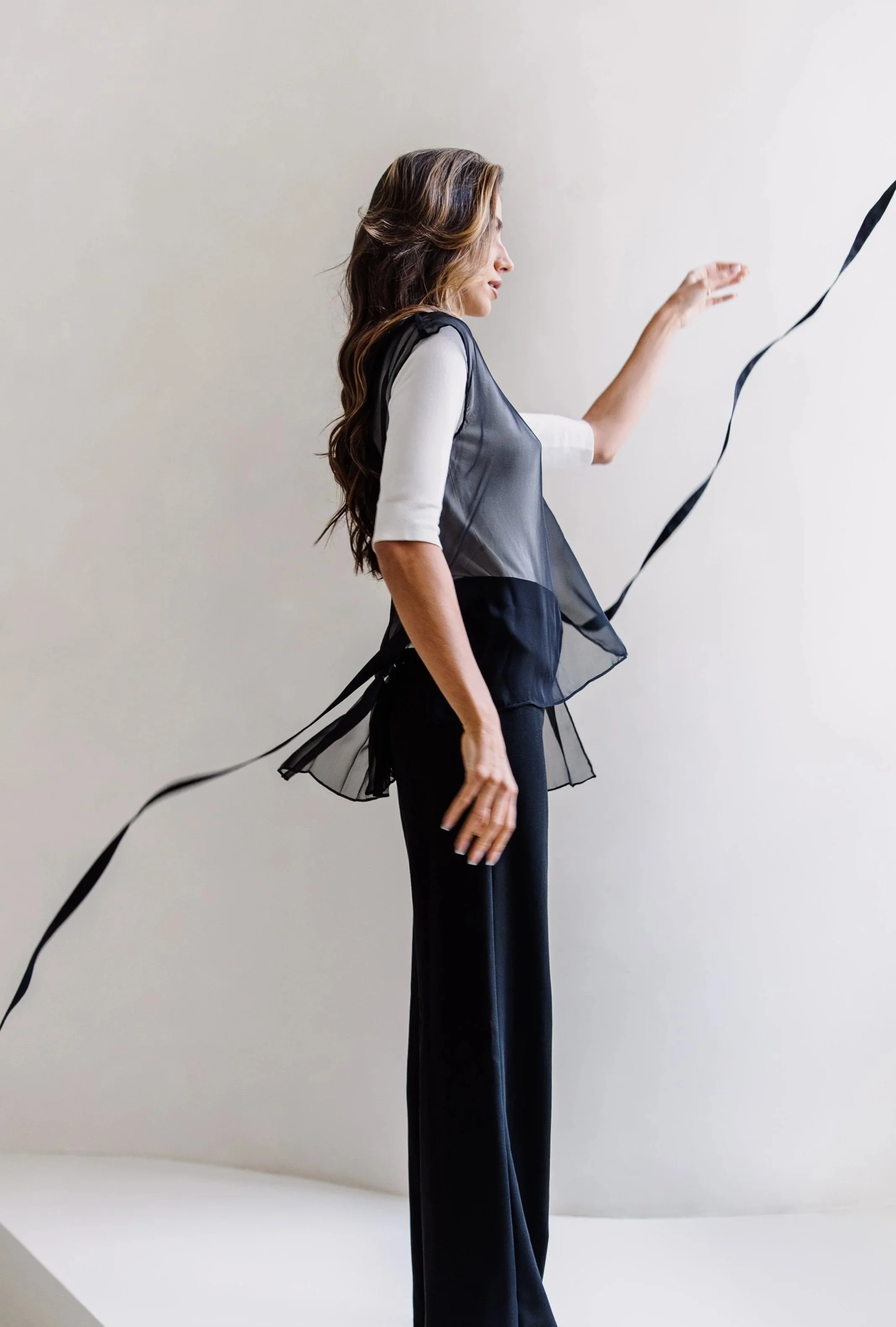 Woman with long wavy hair, wearing a white top with black sheer overlay and black pants, standing sideways in front of a plain light-colored background.