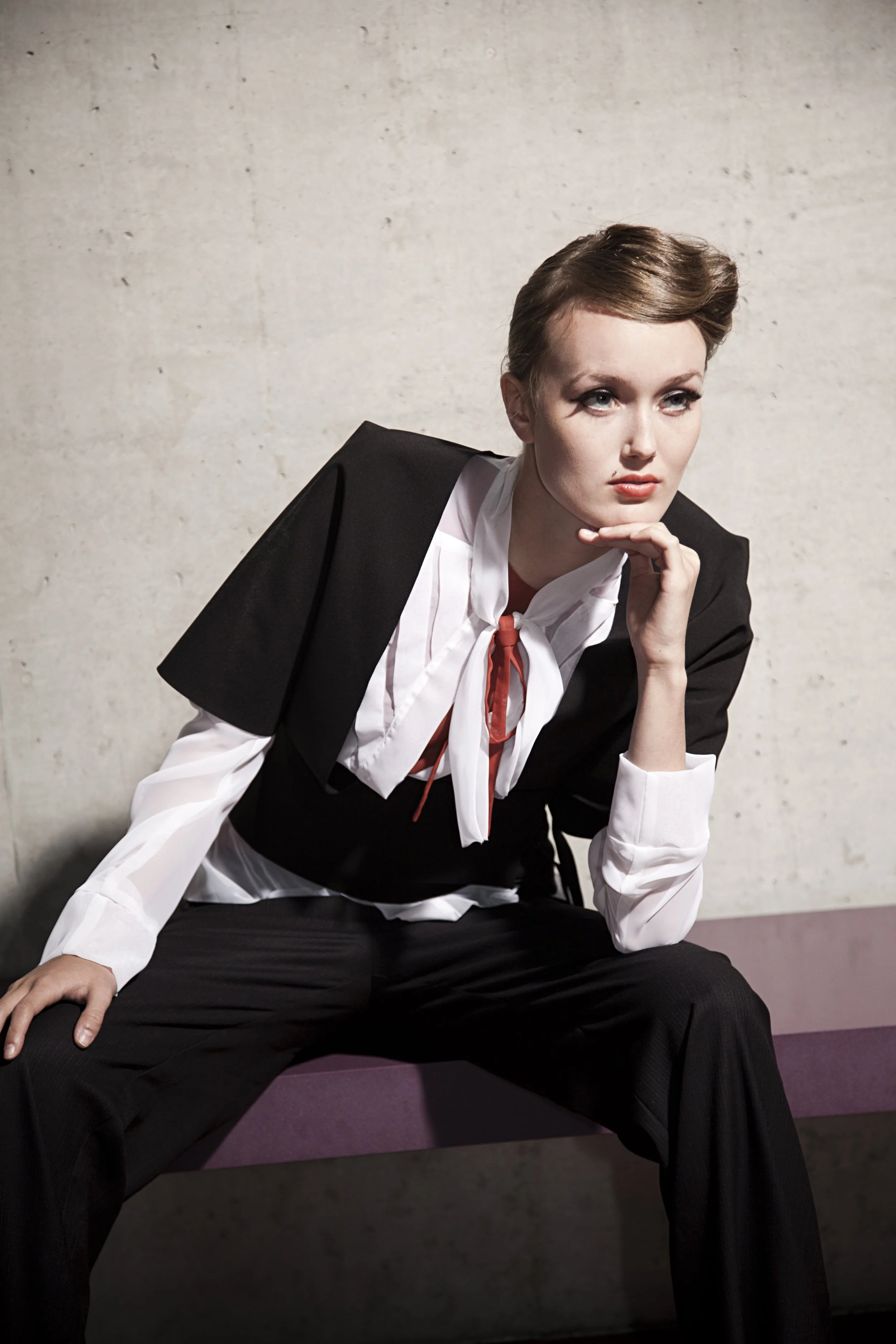 Žena v černém blejzru a bílé košili s červenou kravatou sedí na růžové lavičce u betonové zdi a opírá si bradu o ruku.