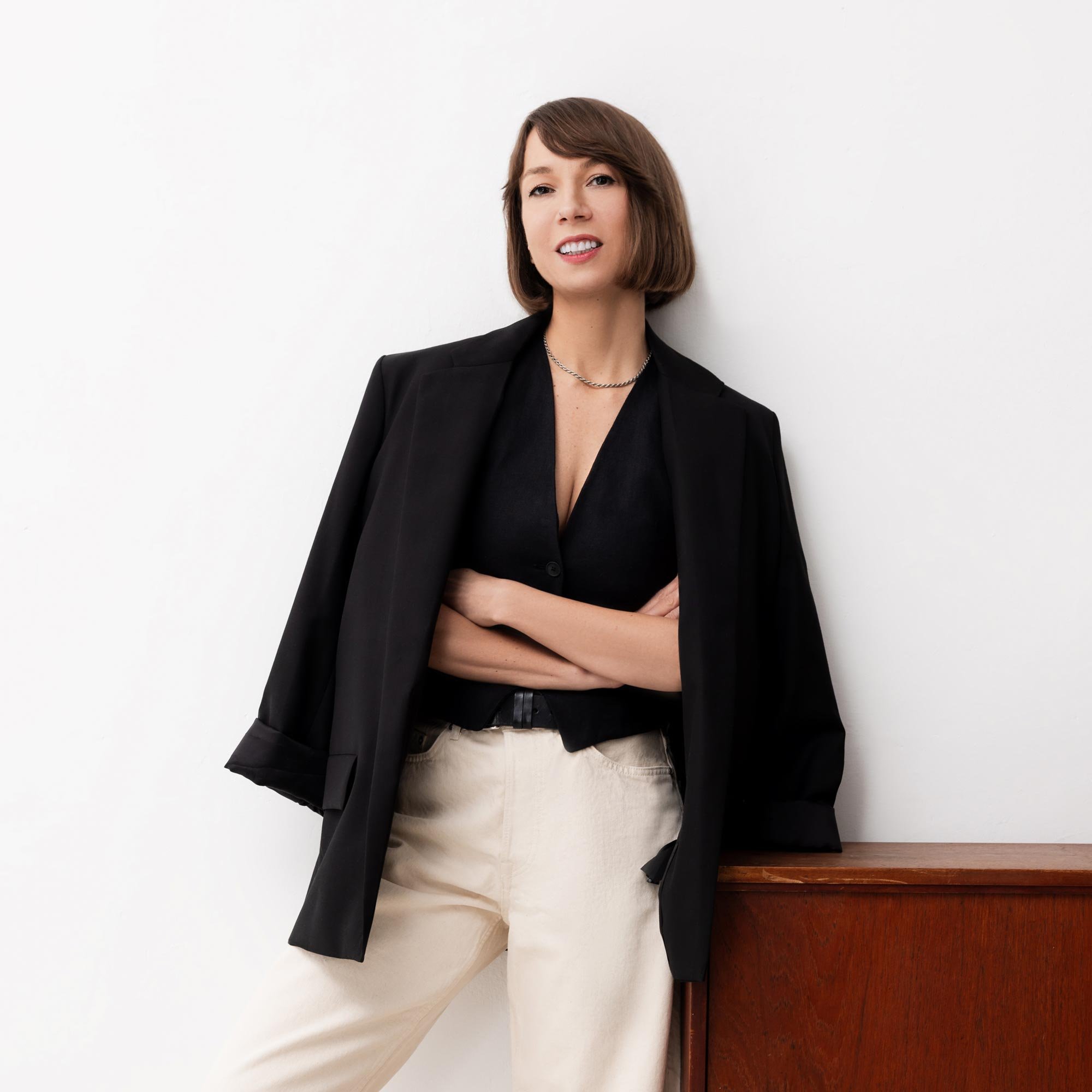 Woman with short brown hair wearing a black blazer over a black top and beige pants, standing with arms crossed against a white wall with a wooden surface nearby.