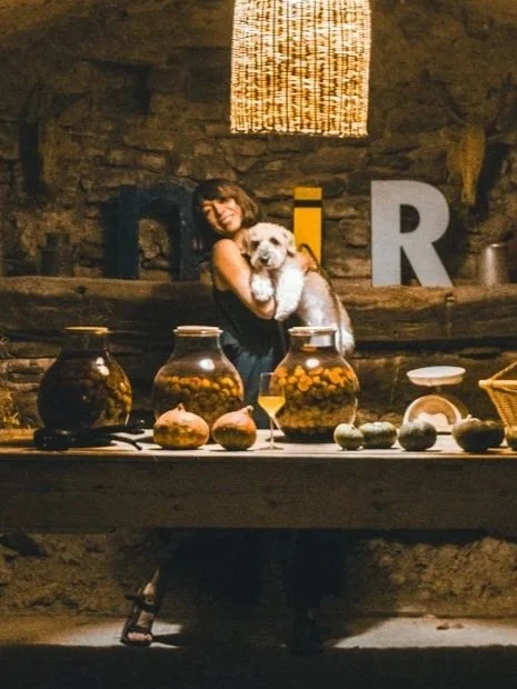A woman holding a small white dog, standing behind a table with jars of pickles and other items, in a rustic setting with large letters 'D' and 'R' in the background.