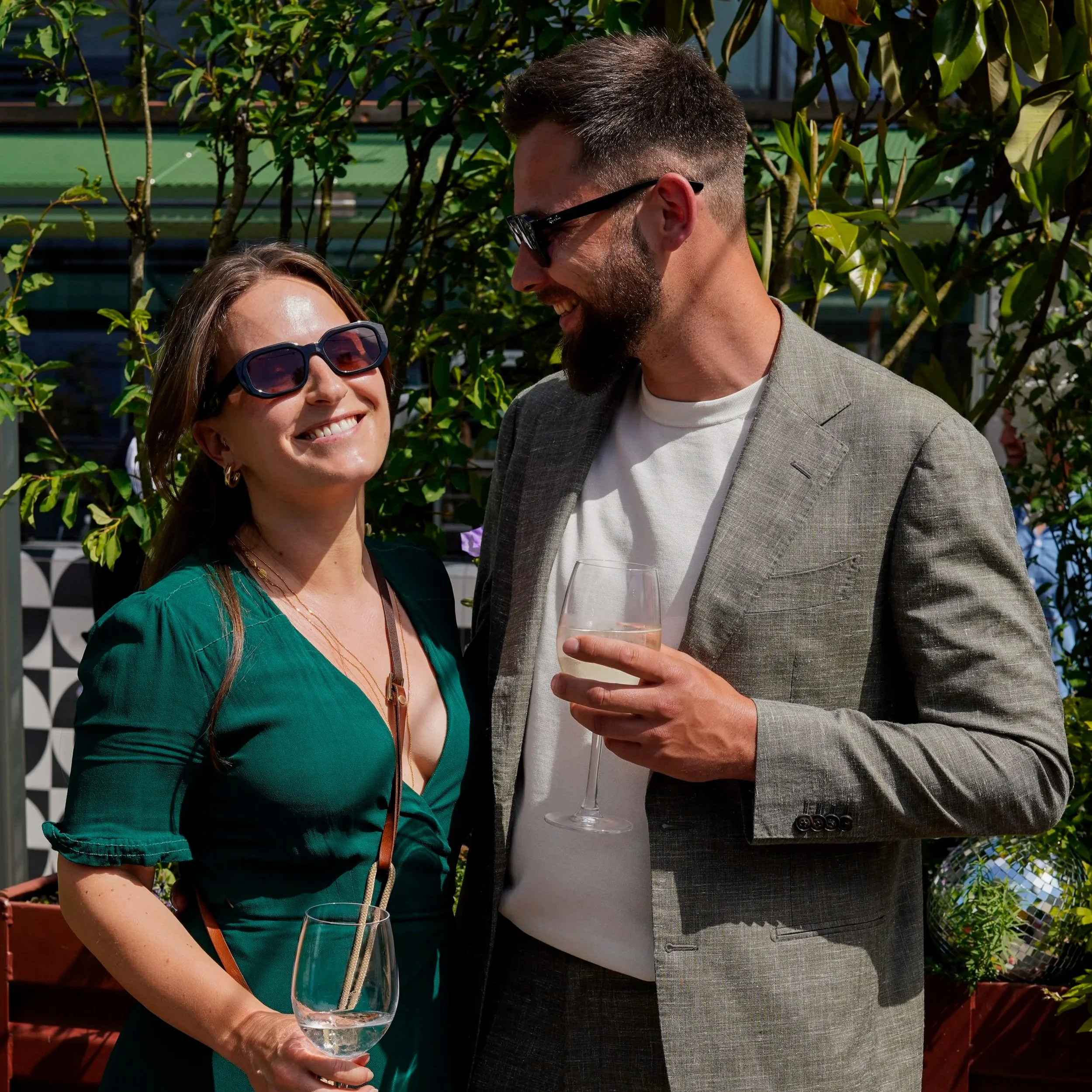 Ein Mann und eine Frau, die bei einer Party im Freien lachen, mit Gläsern in der Hand, umgeben von Grünpflanzen und Dekorationen.