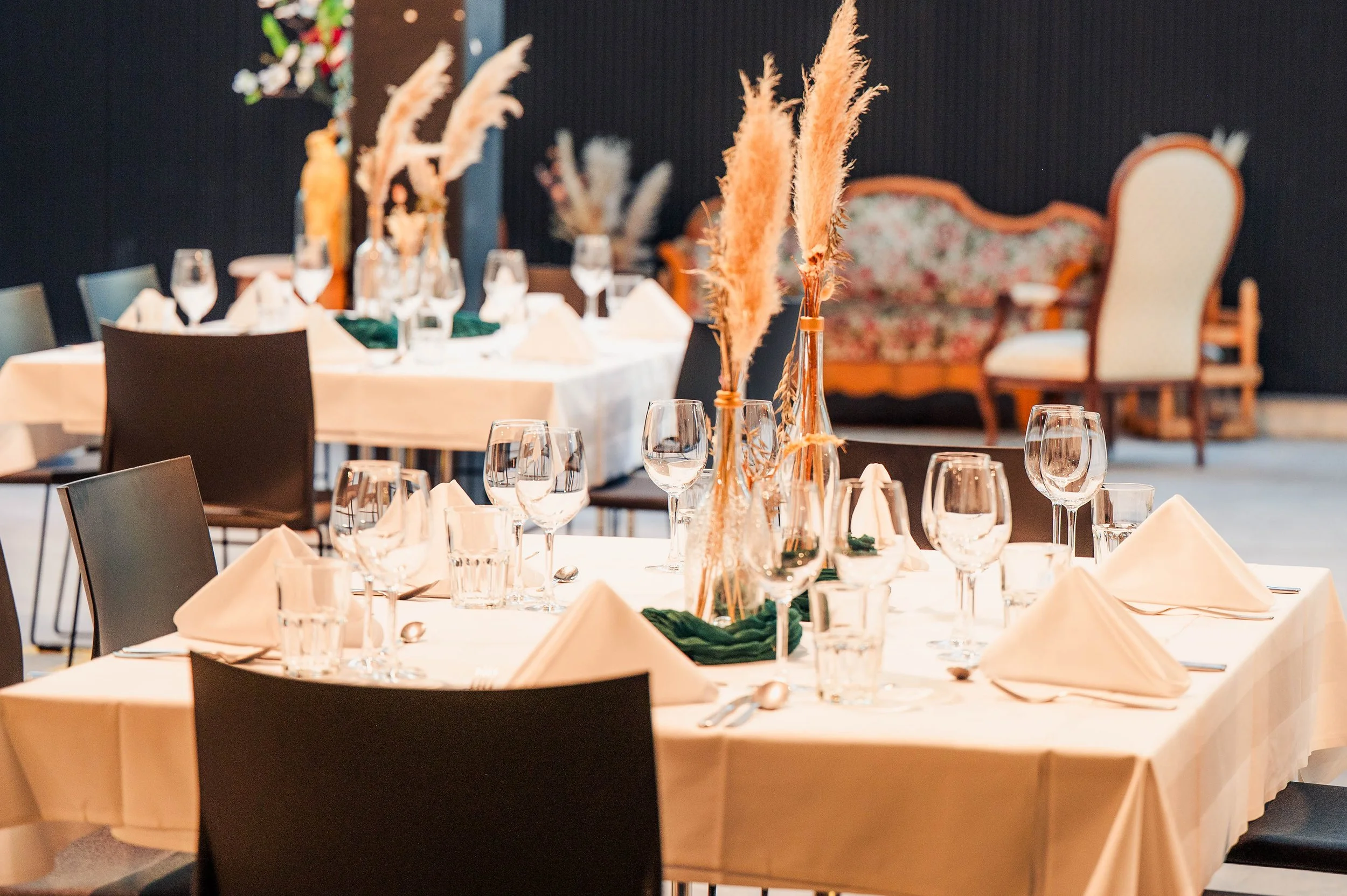 Ein festlich gedeckter runder Tisch mit weißen Tischtüchern, mehreren Weingläsern, Wasserbechern, weißen Stoffservietten und silbernen Besteckteilen, dekoriert mit hohen Gläsern mit Pampasgras als Mittelstücke, in einem eleganten Raum mit dunklem Hin