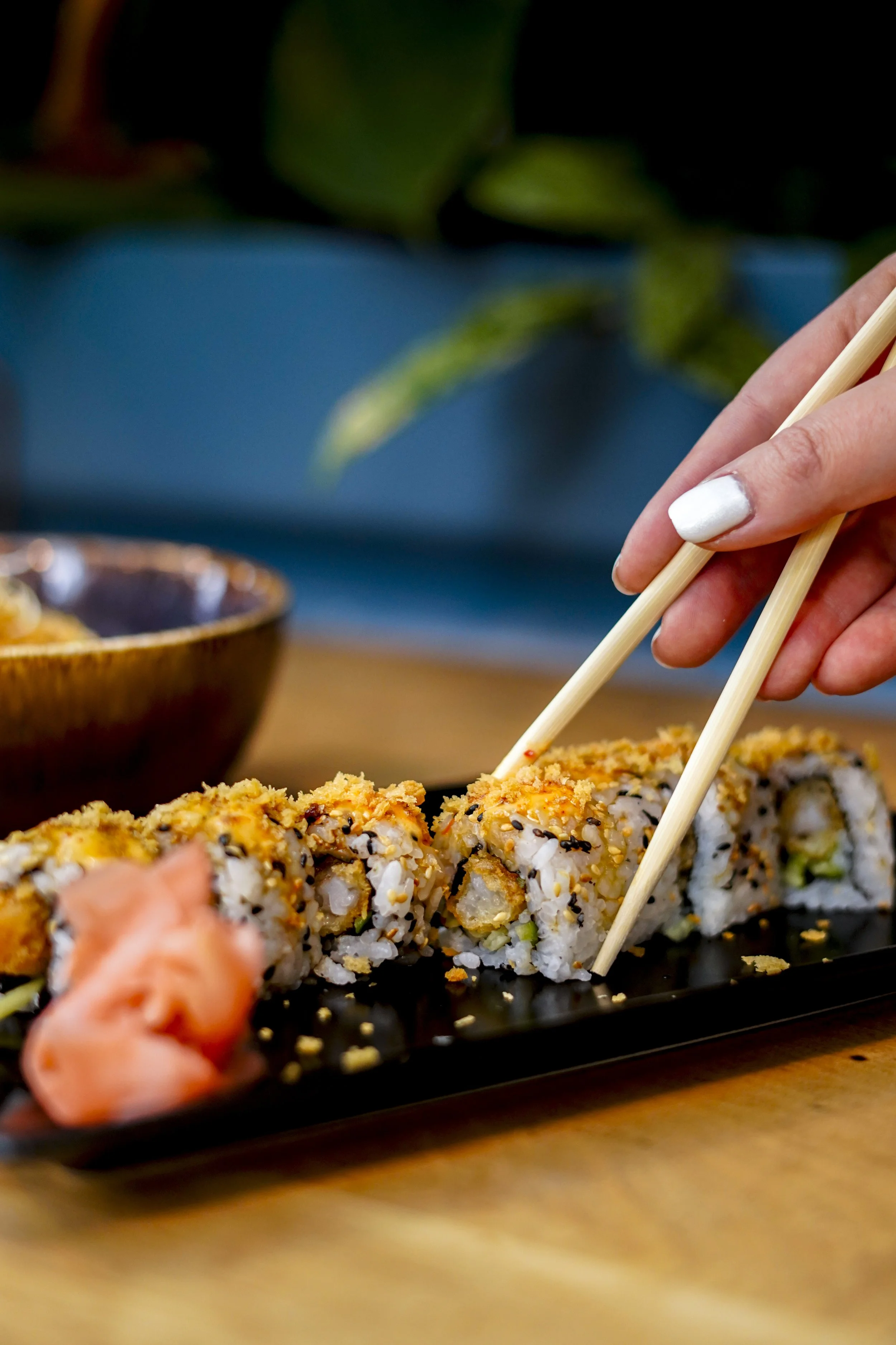 Nahaufnahme von Sushi-Rollen mit Händen, die mit Essstäbchen greifen, auf einem schwarzen Teller mit eingelegtem Ingwer