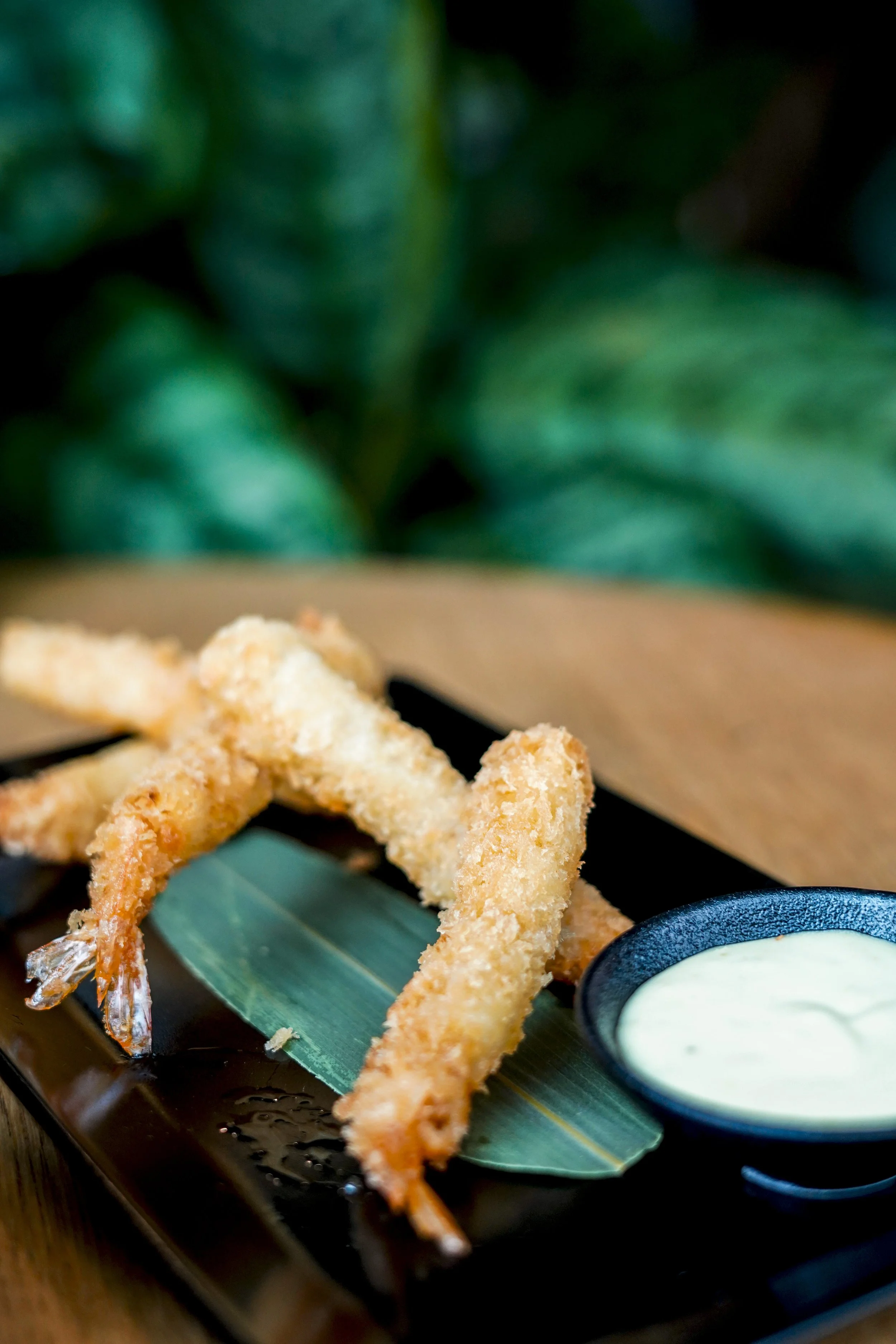 Frittierte Garnelen mit grüner Dip-Sauce auf einem schwarzen Teller, Serviert auf einem Holztablett mit grünen Blättern im Hintergrund.