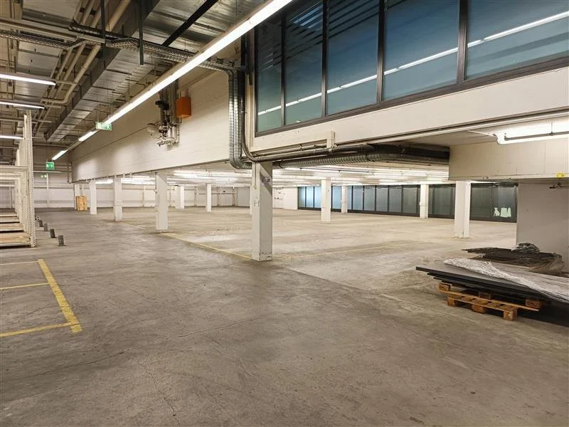 Leerer Parkhaus mit weißen Säulen, grauem Boden, angehängtem Büro oder Technikbereich im zweiten Stock, und einigen Paletten sowie Baumaterialien auf der rechten Seite.