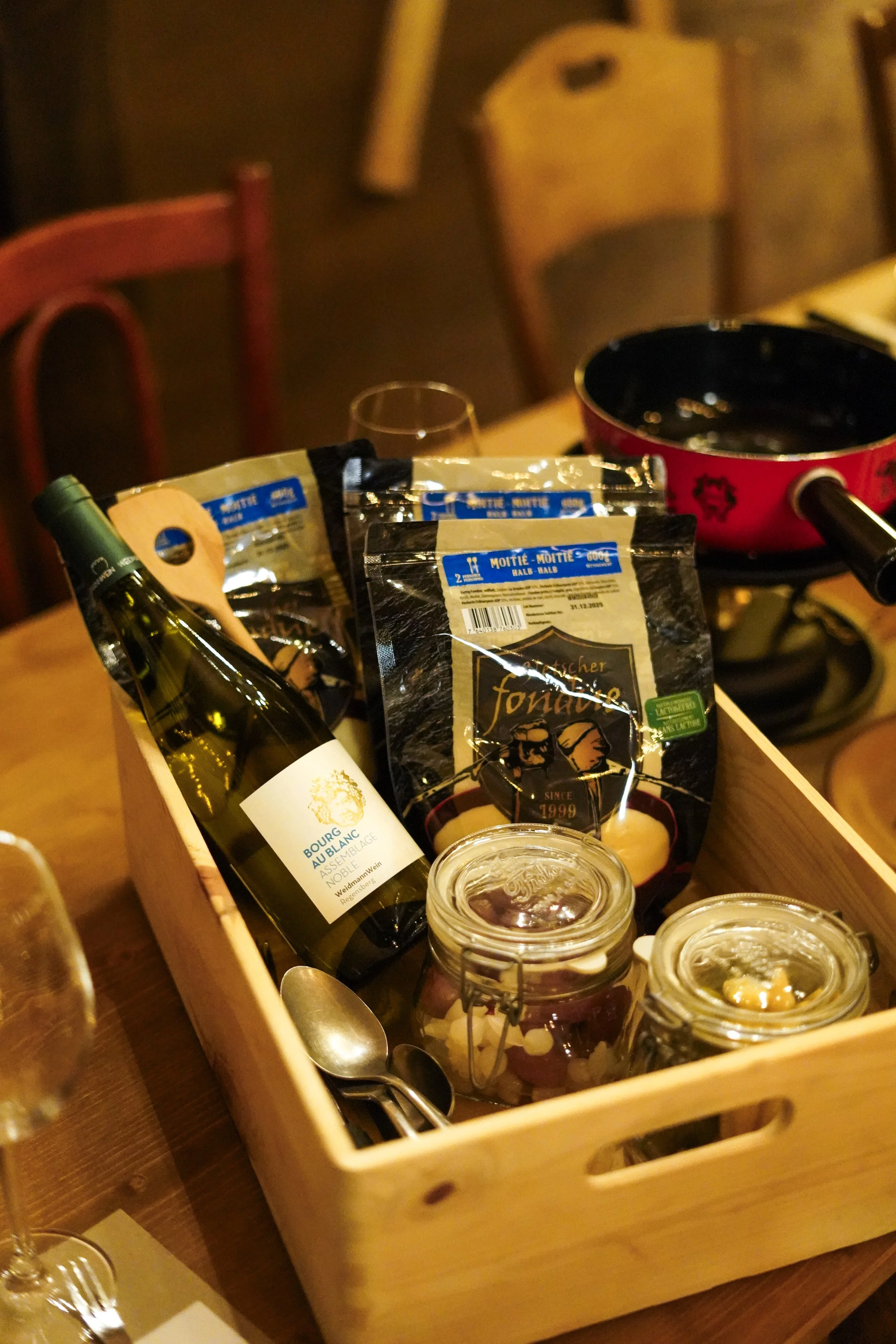 Holzkiste mit Weinflasche, Schalen, Gläsern, Besteck, Süßigkeiten und einer Pfanne auf einem Tisch.