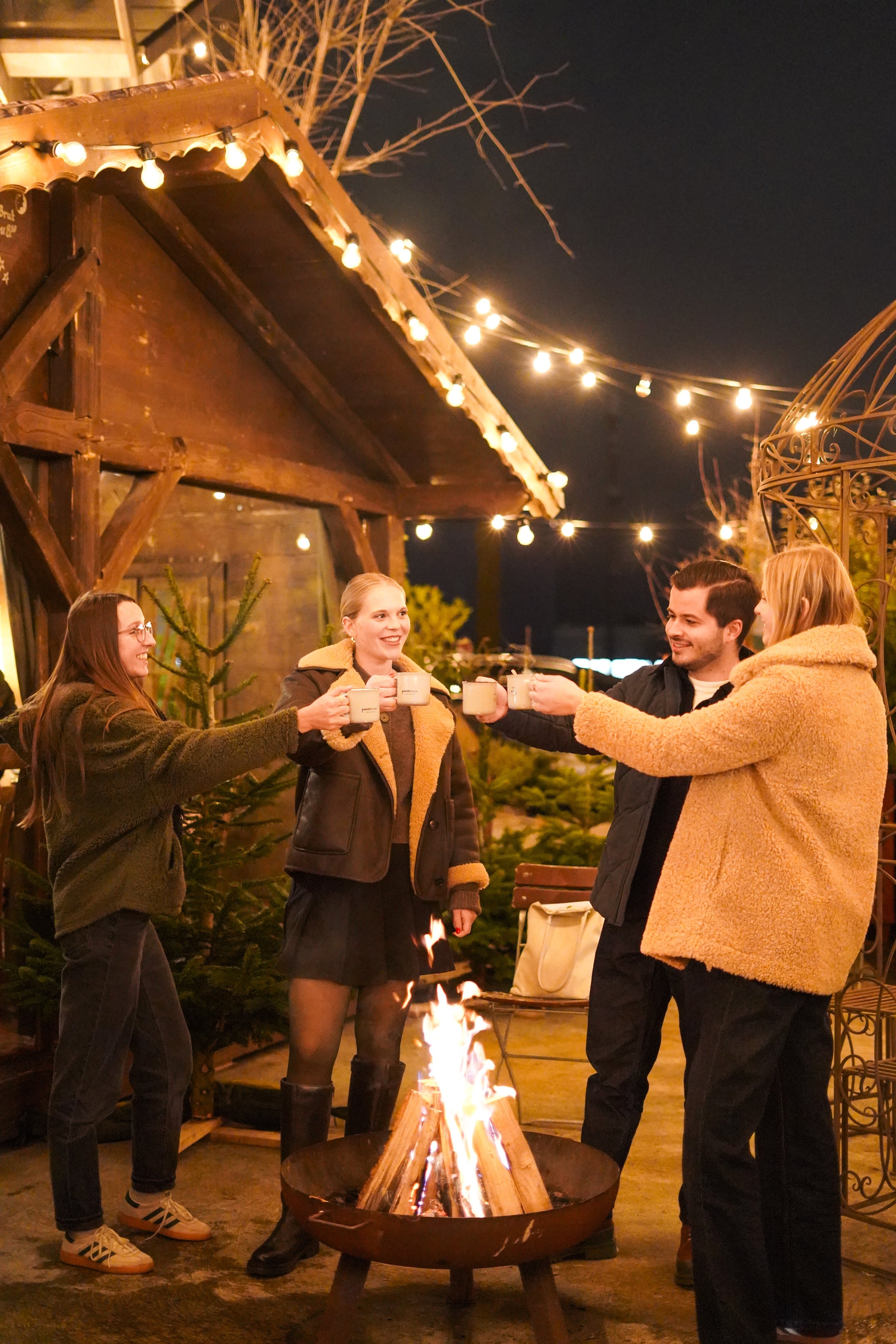 Vier Menschen stoßen mit Tassen an einem Lagerfeuer im Garten bei Nacht an, umgeben von Lichterketten und Weihnachtsbäumen.