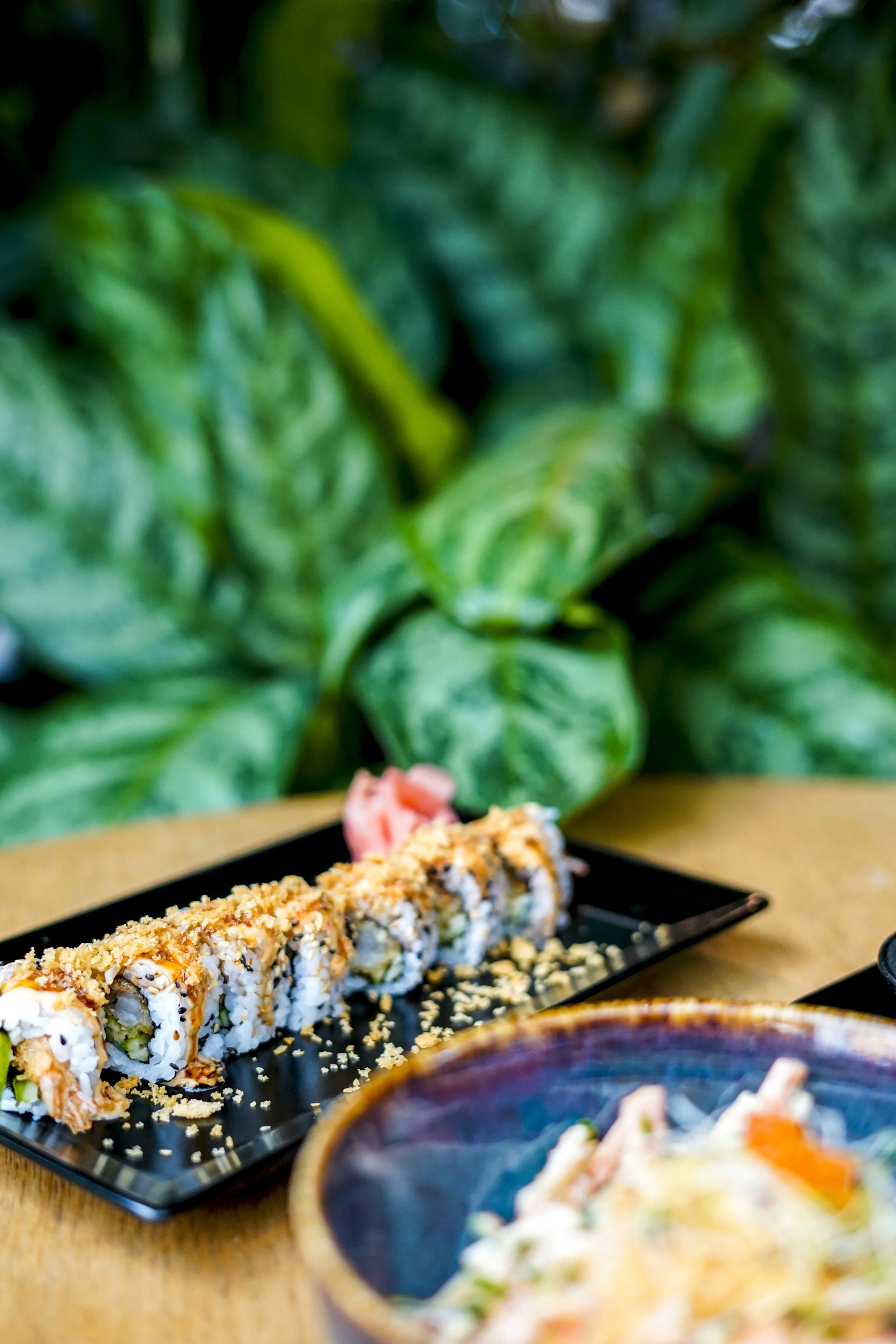 Sushi rollen auf einem schwarzen Teller mit gepresstem Wasabi und Ingwer, im Vordergrund eine Schüssel mit zusätzlichem Sushi, im Hintergrund grüne Blätter.