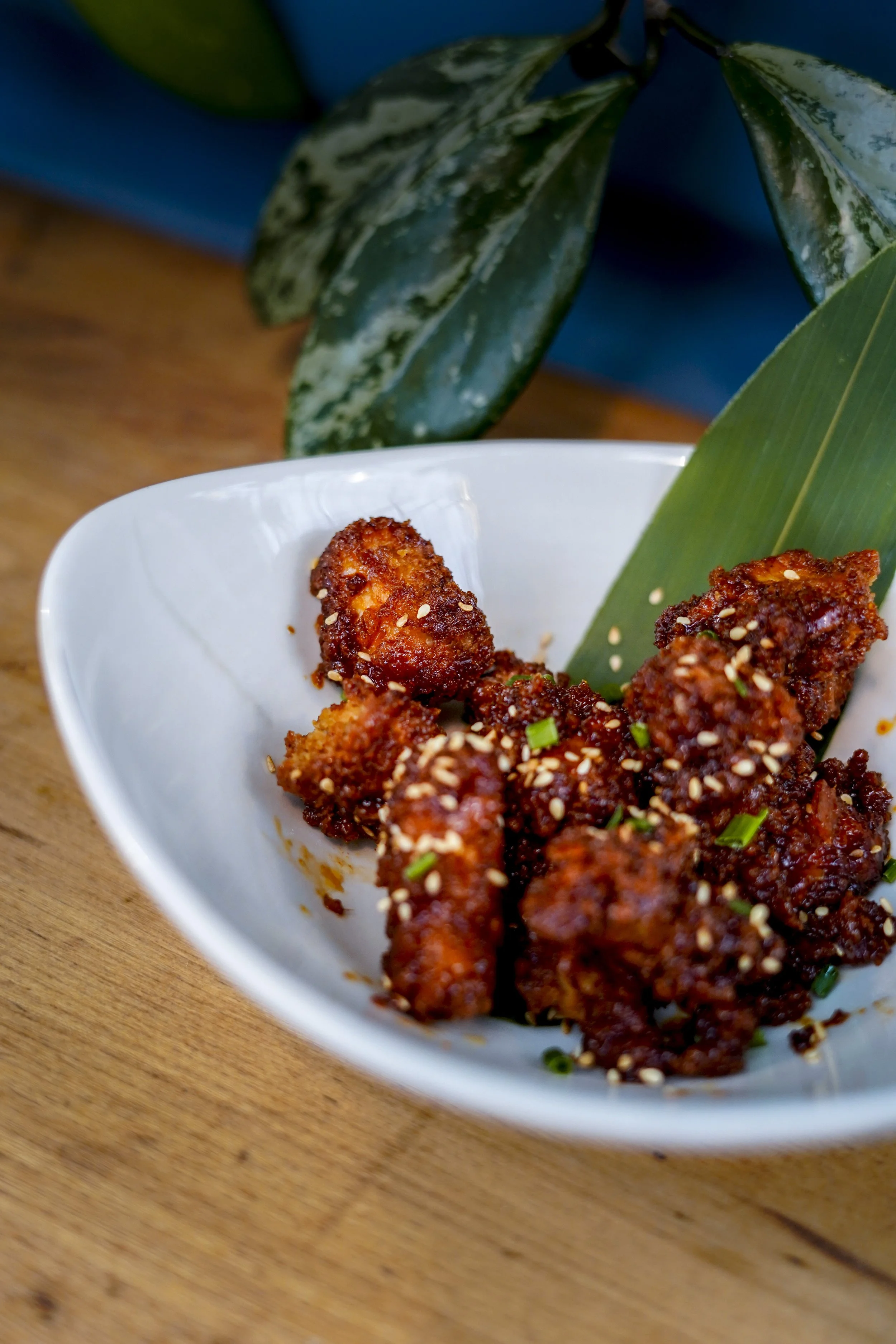 Knusprige asiatische Chicken Wings in einer weißen Schale mit Sesamsamen, Zwiebel und grünen Frühlingszwiebeln, auf einem Holztisch mit grünem Blatt und Pflanze im Hintergrund.